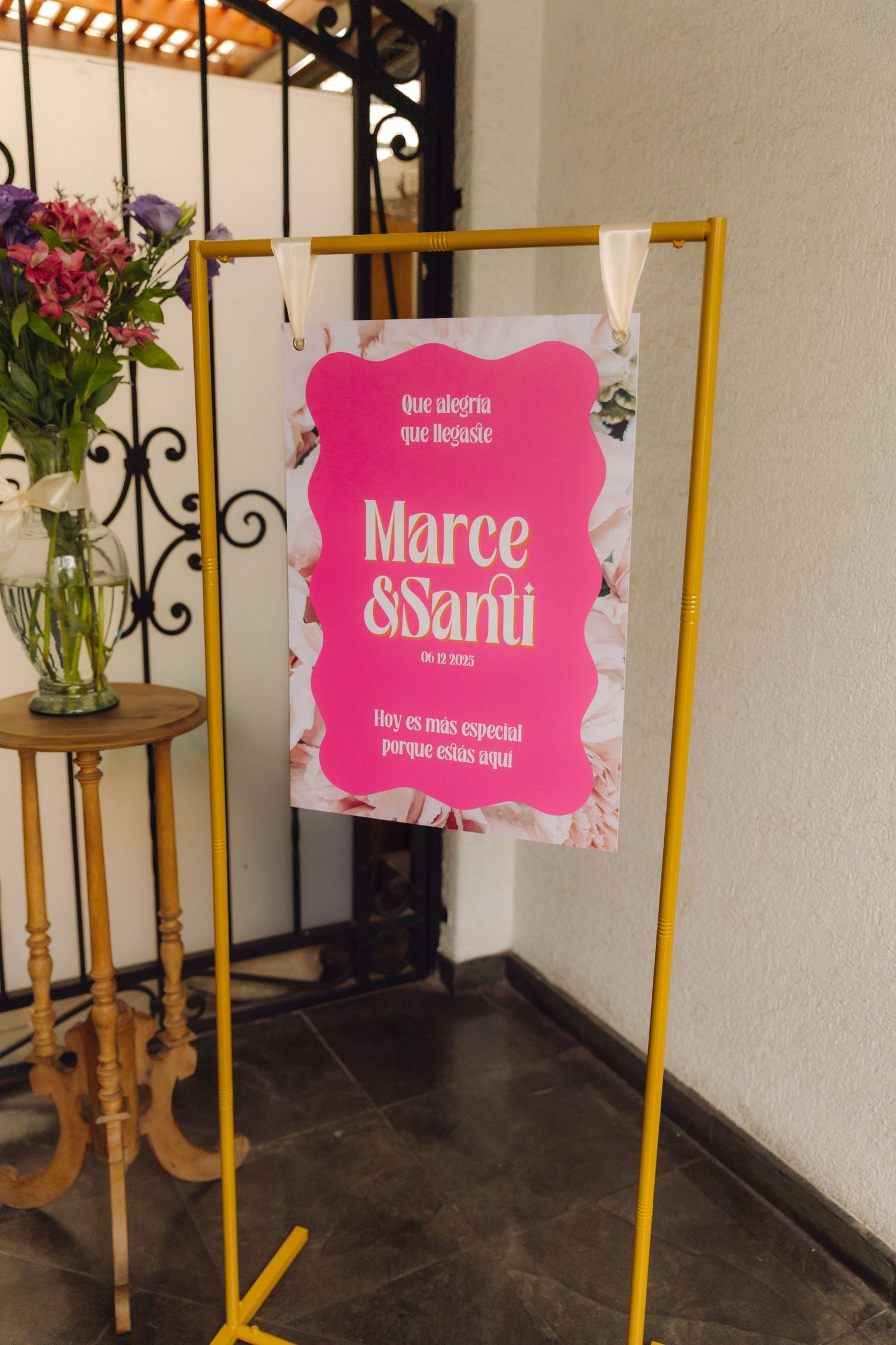 Pink and white wedding welcome sign for Marce and Santi displayed on a yellow stand with flowers.
