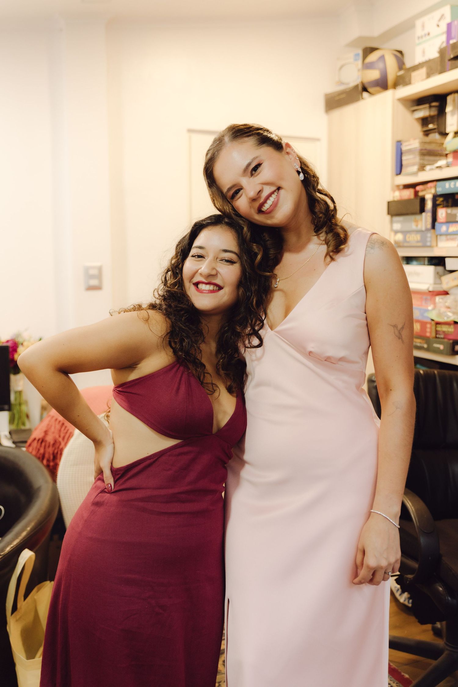Two smiling women in elegant formal dresses pose together at a wedding preparation salon.