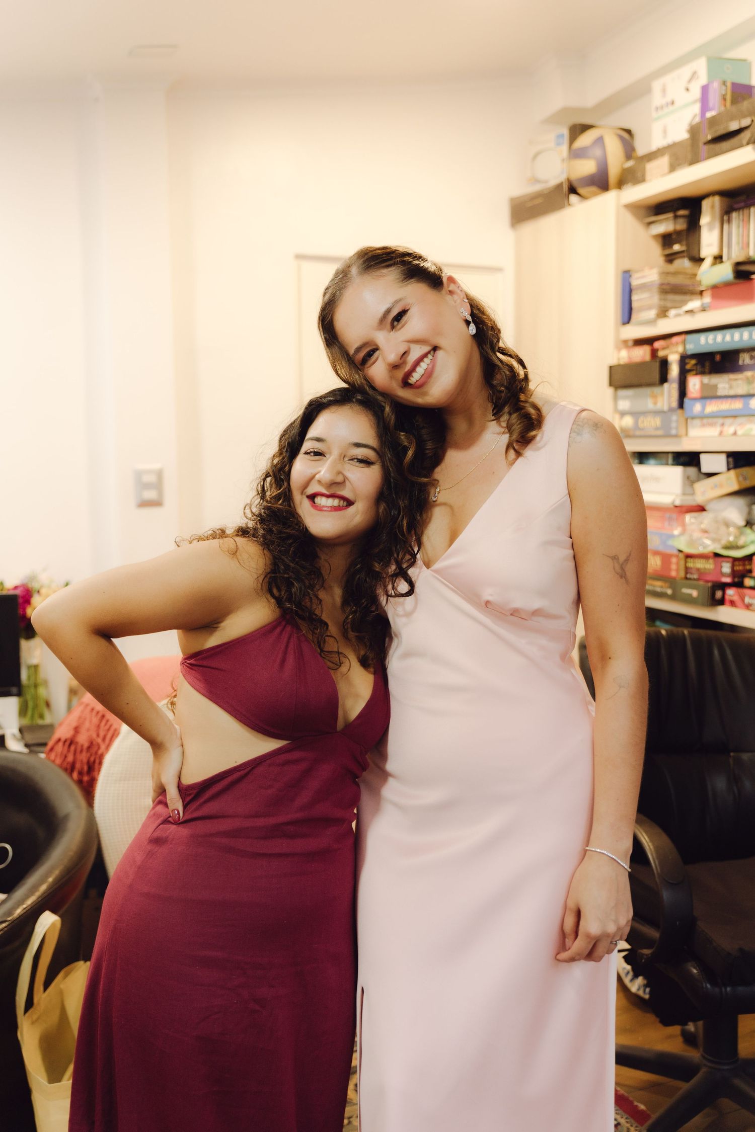 Two young women in formal gowns smile and embrace in a dressing room before a wedding event.