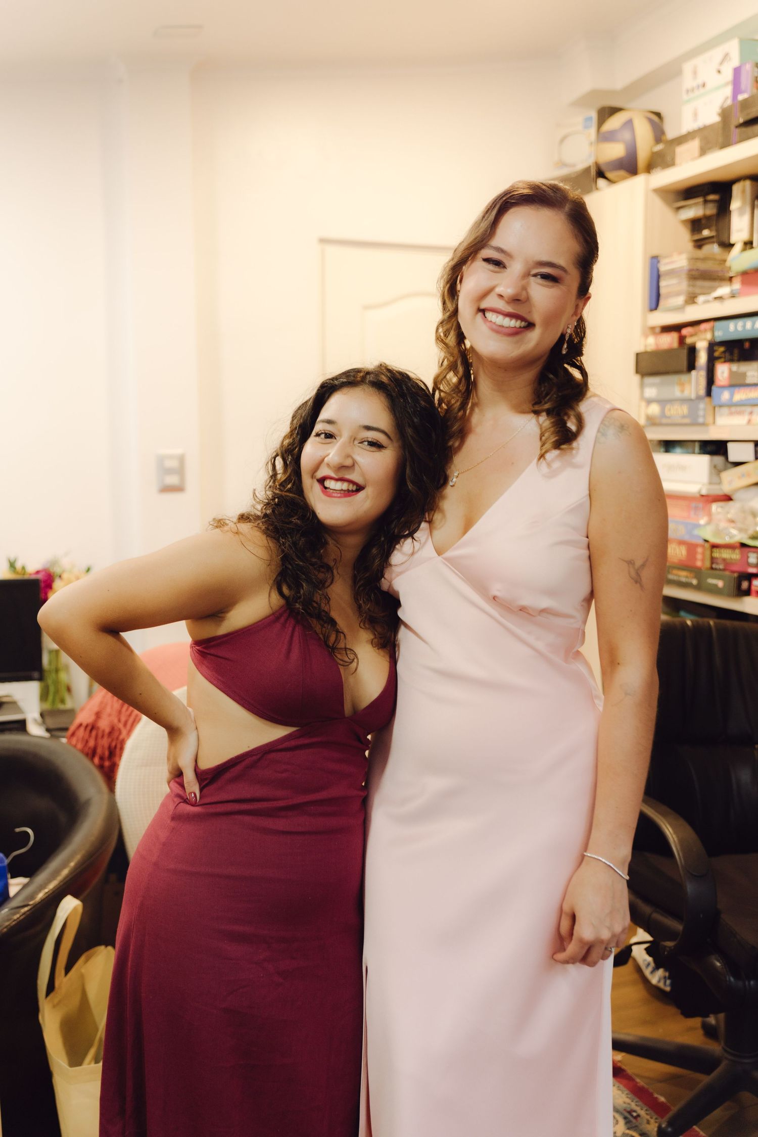 Two happy women in formal dresses pose together smiling in a bridal preparation room.