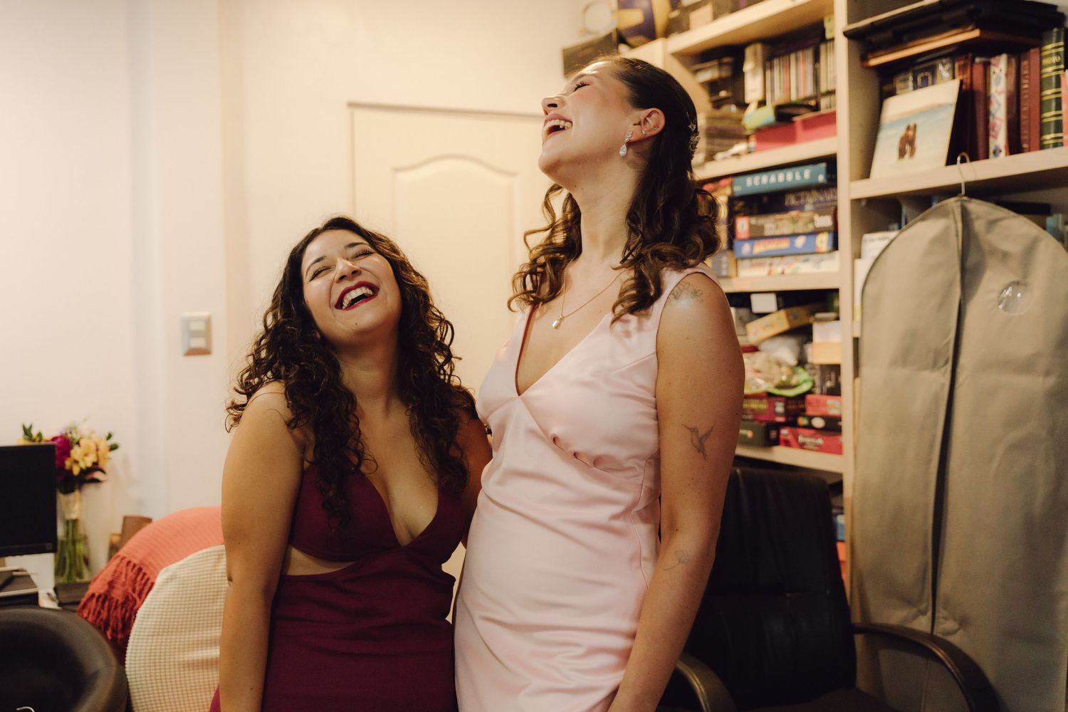 Two women in formal dresses laugh joyfully together in a room while getting ready for a wedding.