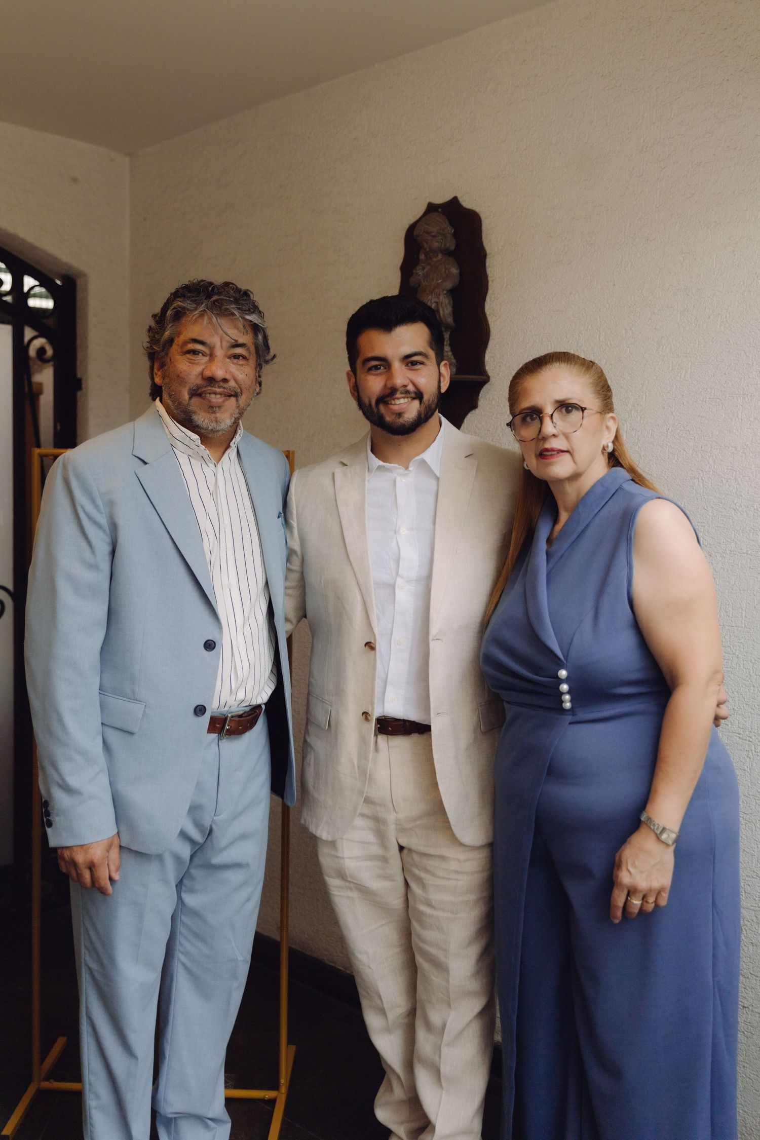 Young man in beige suit poses with his parents dressed in blue formal attire at a wedding.