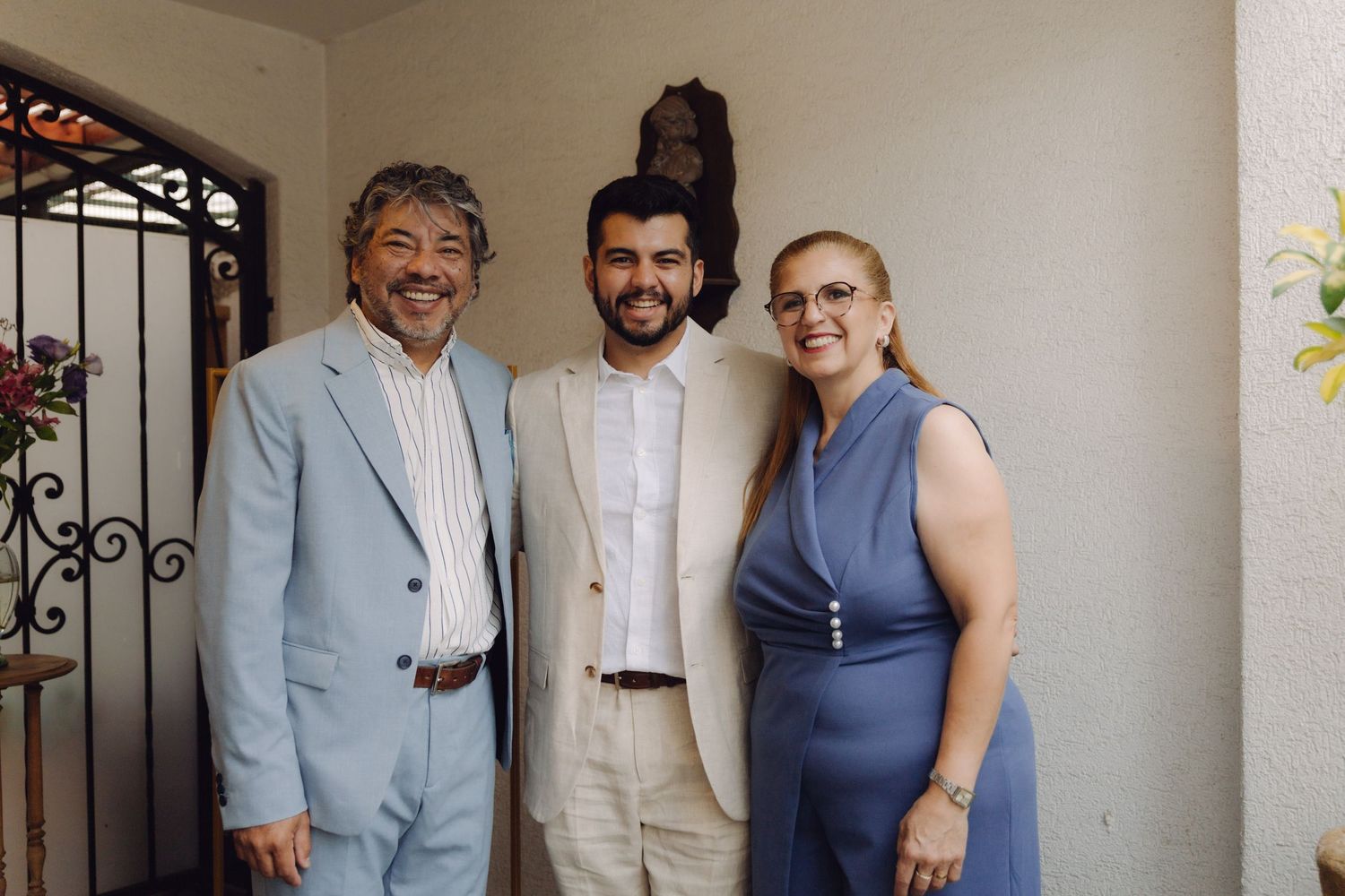 Smiling family trio with parents in blue formal wear and son in beige suit at a wedding celebration.