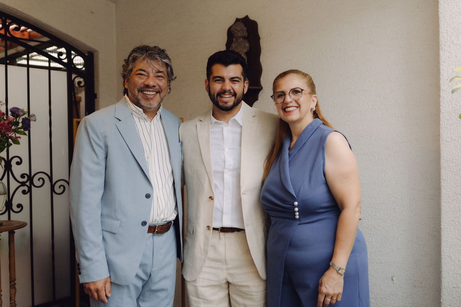 Family of three posing outdoors, son in beige suit flanked by parents in elegant formal attire.