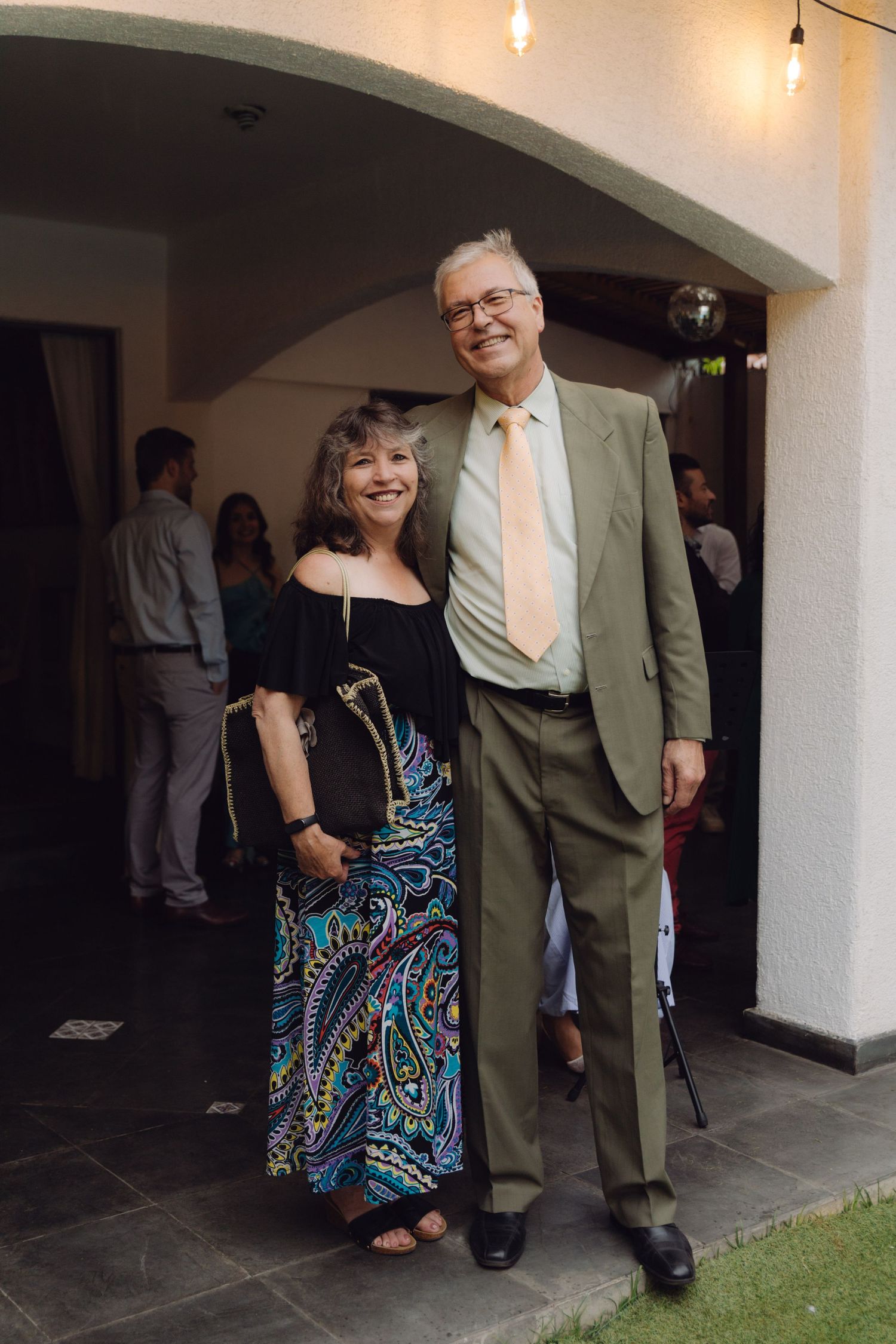 Elegant older couple posing together at a formal event, man in olive suit with tie, woman in colorful paisley skirt.