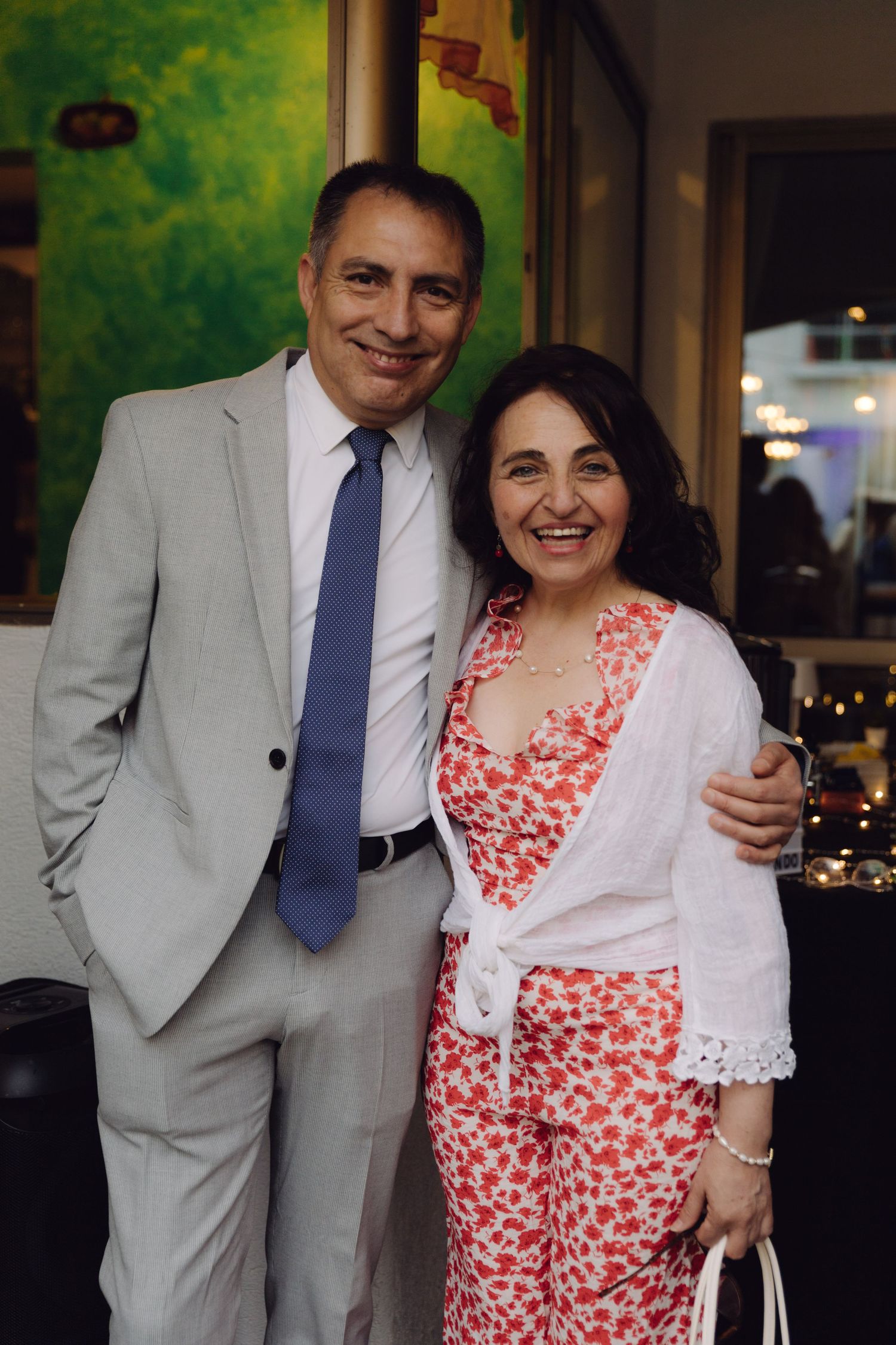 Smiling couple posing together at an indoor event, man in gray suit with blue tie, woman in floral outfit.