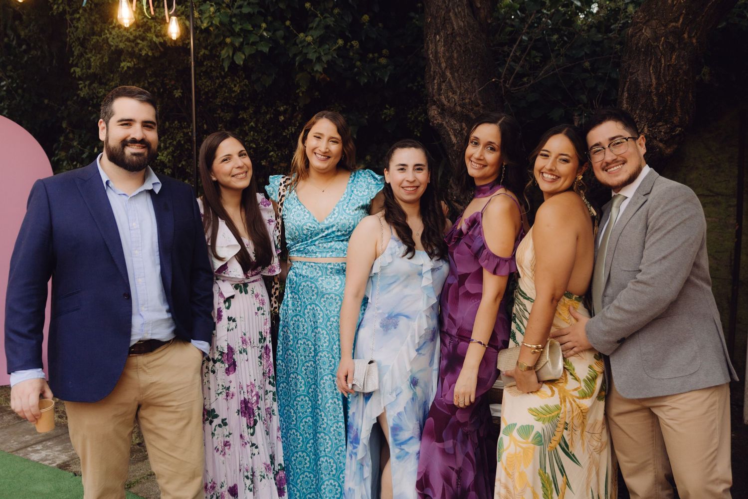 Group of seven friends posing together outdoors at a garden party wearing elegant formal attire.