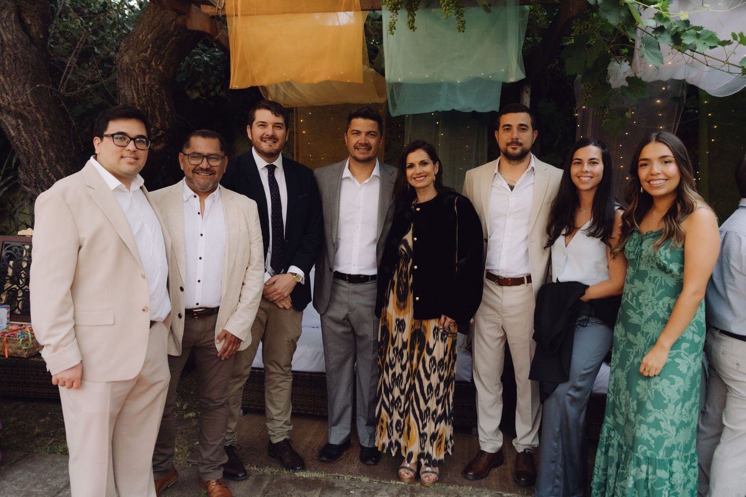 Group of eight elegantly dressed guests posing together outdoors under trees at a formal celebration.