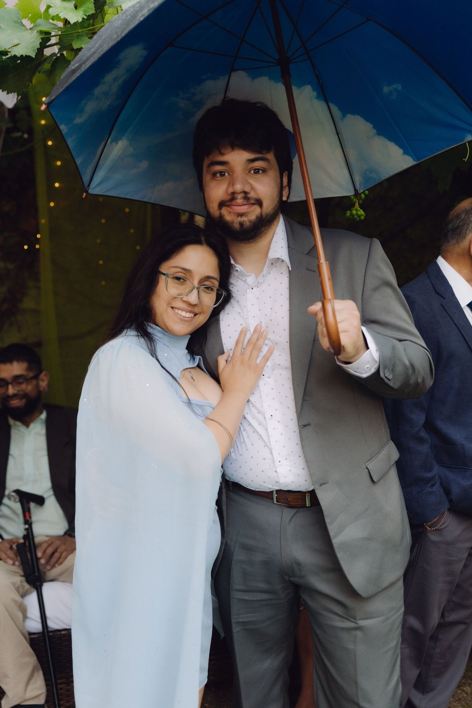 Happy couple posing under a decorative blue umbrella at an outdoor wedding ceremony in gray suit.