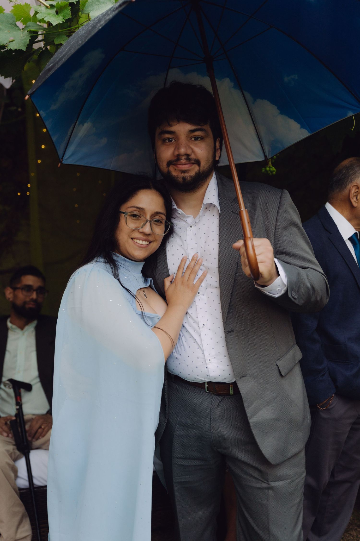 Smiling couple sharing a blue umbrella at an outdoor event, woman in light blue wrap, man in gray suit.