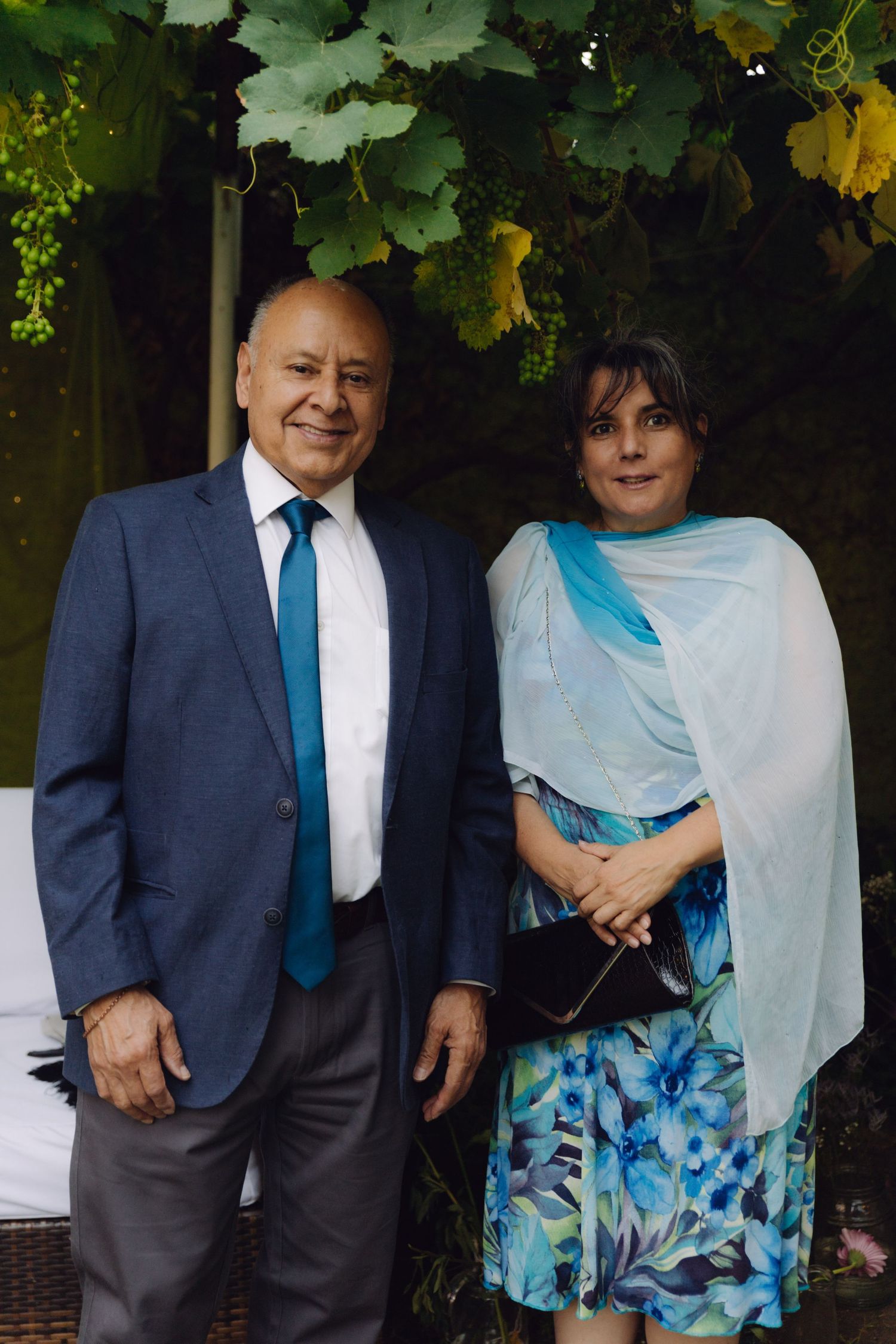 Mature couple posing outdoors under grape vines, man in navy suit with teal tie, woman in floral dress.