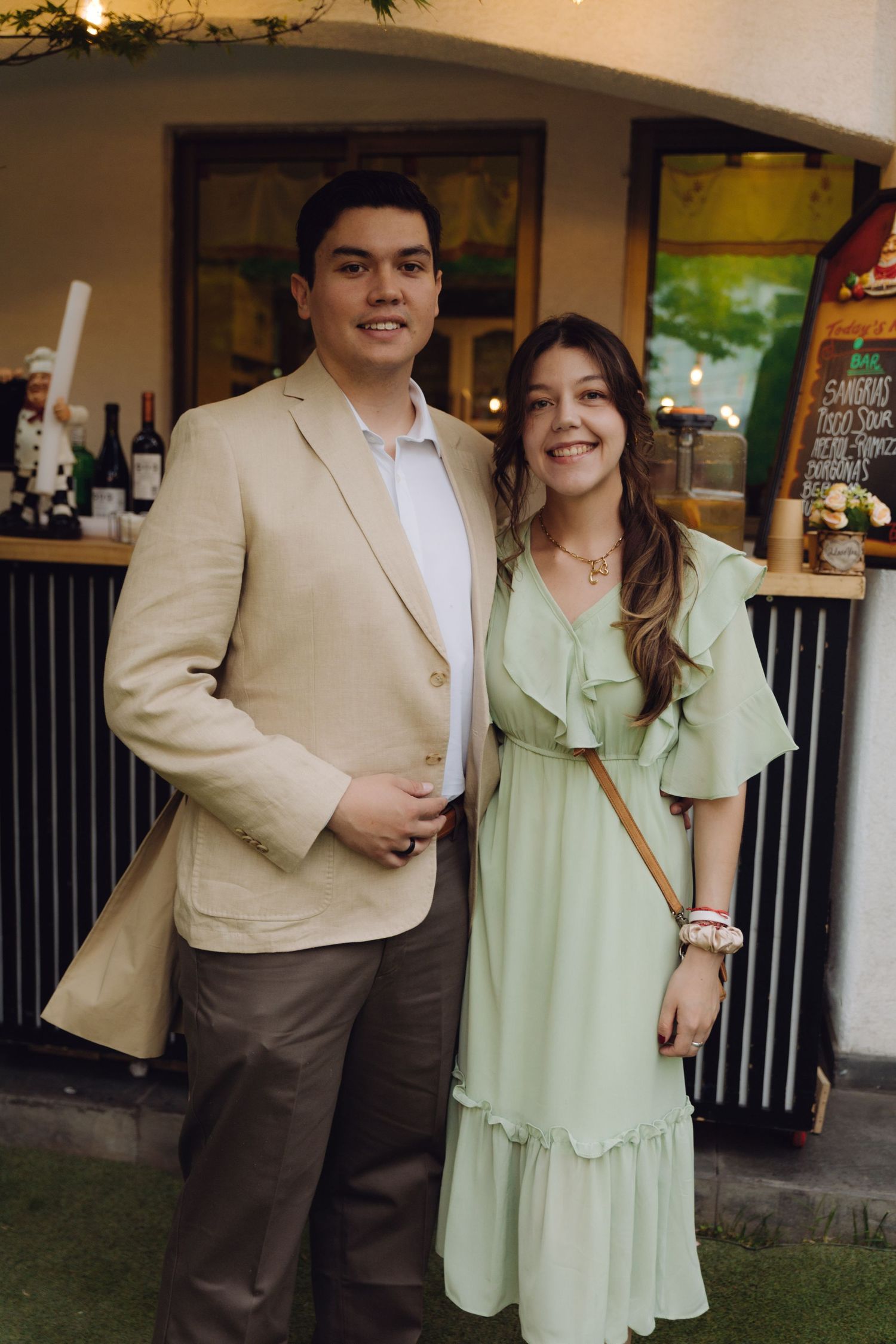 Young couple smiling together at an outdoor event, man in beige blazer, woman in mint green ruffled dress.