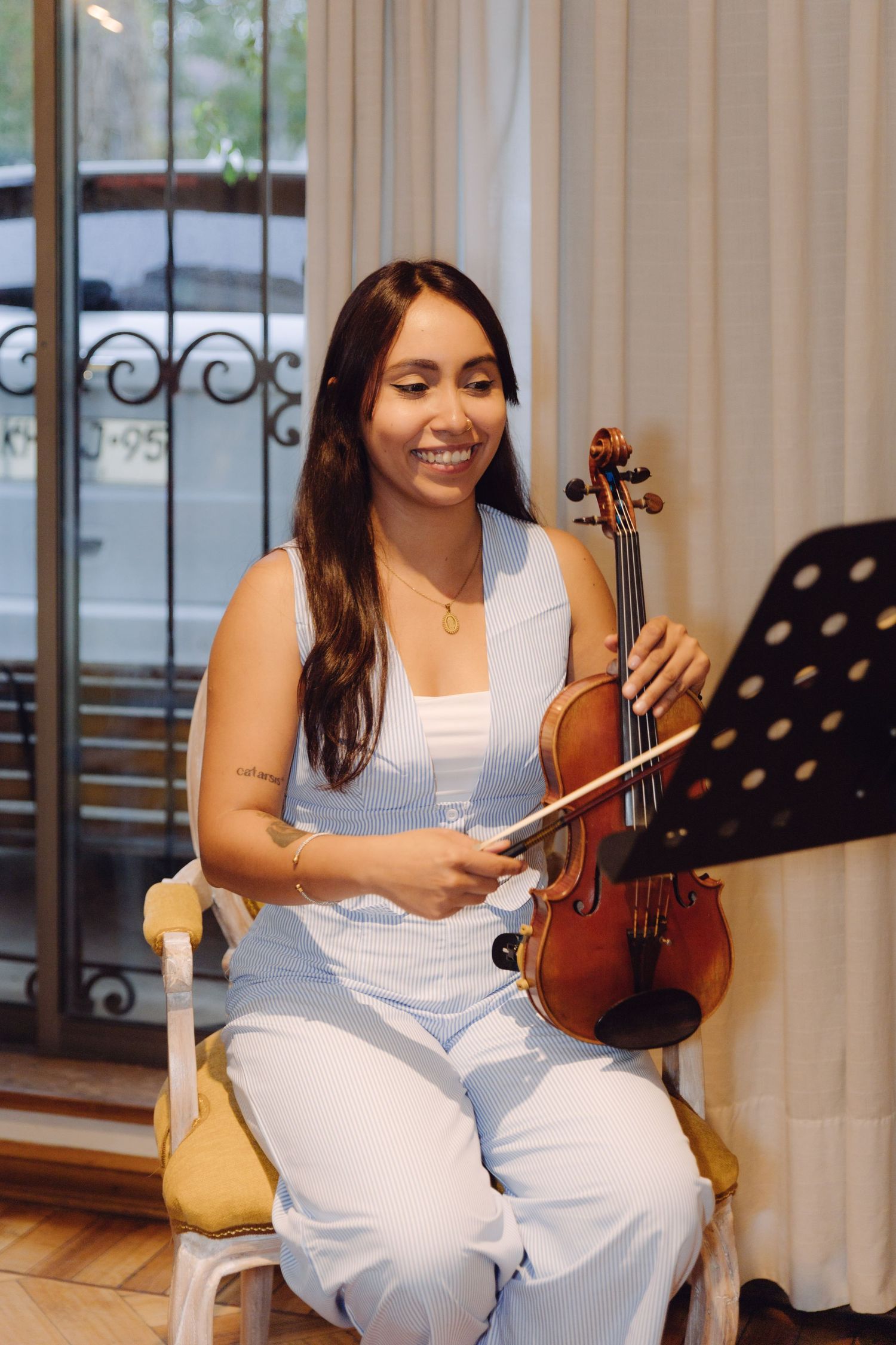 A smiling young woman in white sits holding a violin and bow beside a music stand.