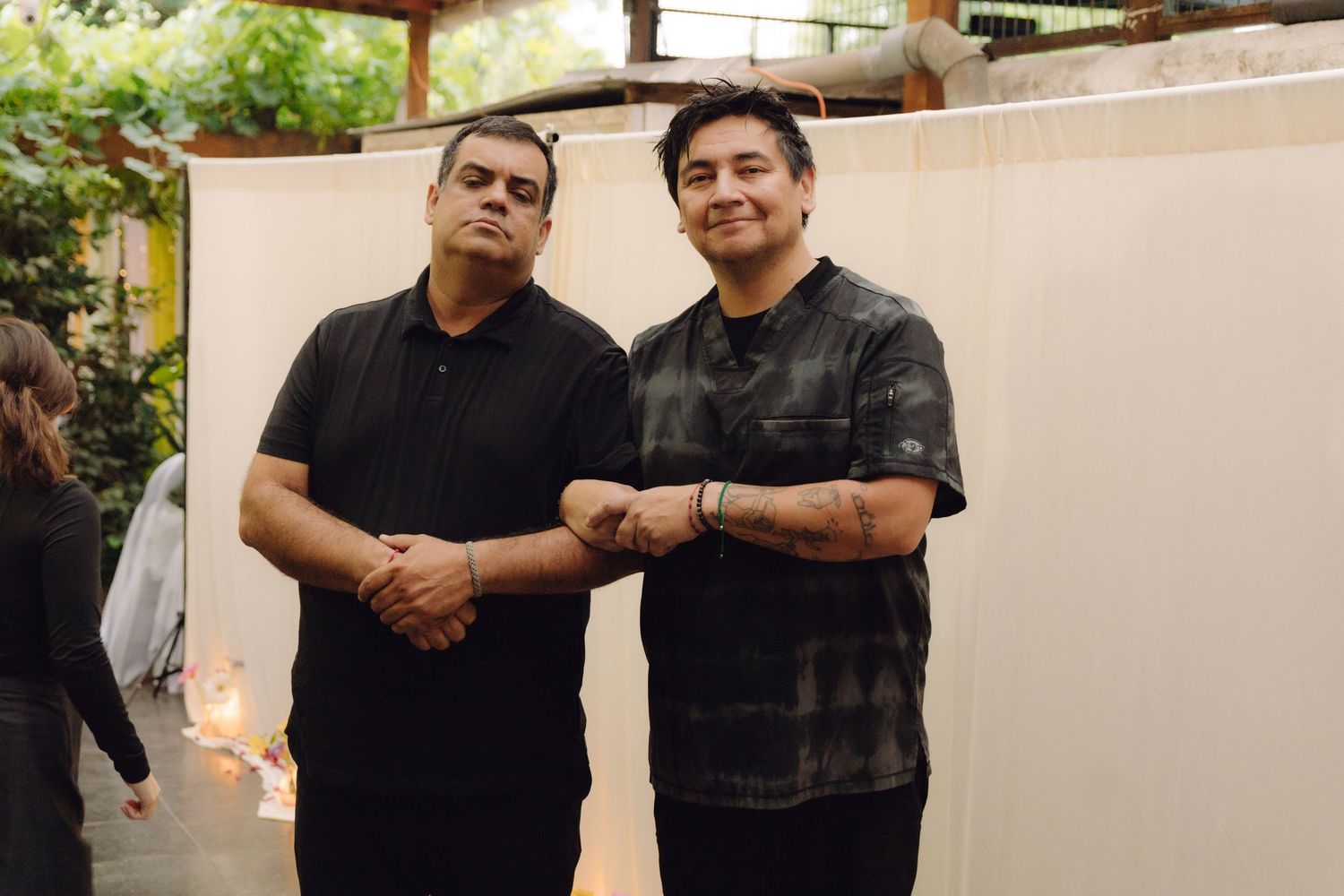 Two men in casual black attire doing a fist bump while posing together at an outdoor venue.