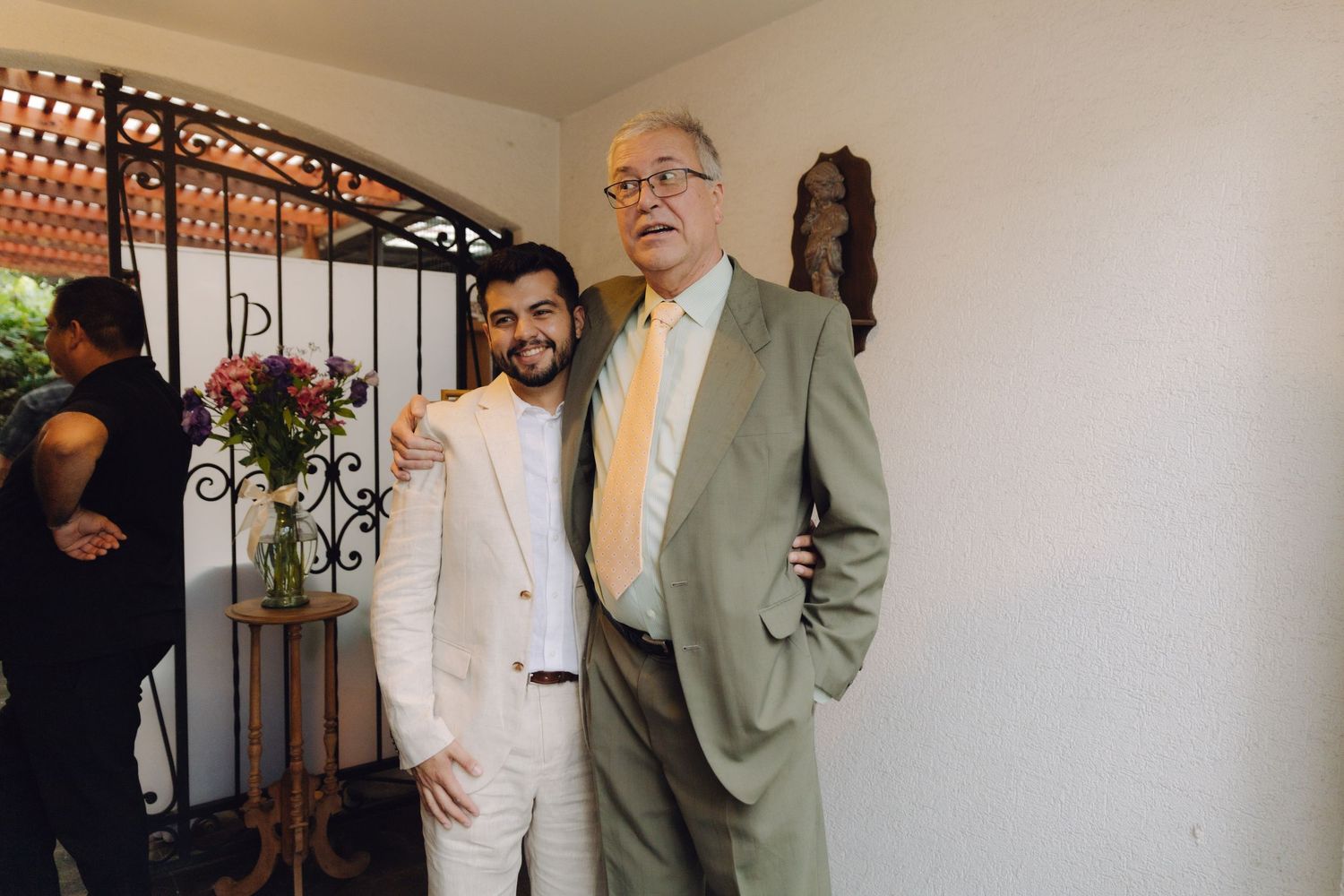 Two men in suits pose together near an ornate iron gate with flowers at a formal event.