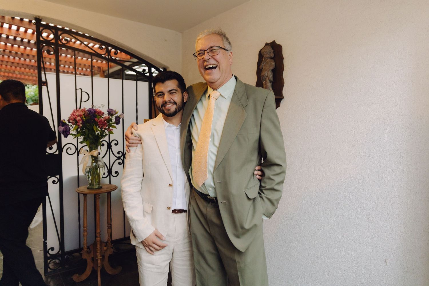Two men in suits laugh together joyfully near a decorative iron gate at a celebration.