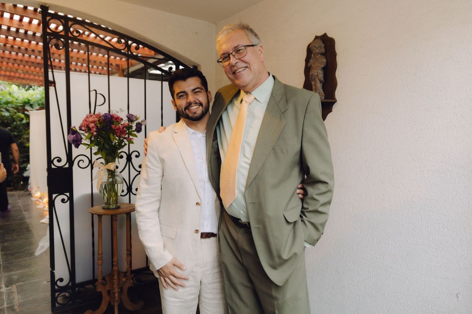 Two men in formal suits smile together outdoors near an ornate black iron gate.