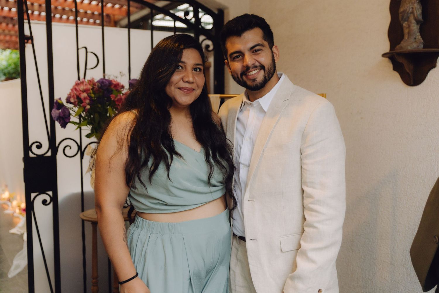 A young couple in formal attire poses together smiling near a decorative iron gate.