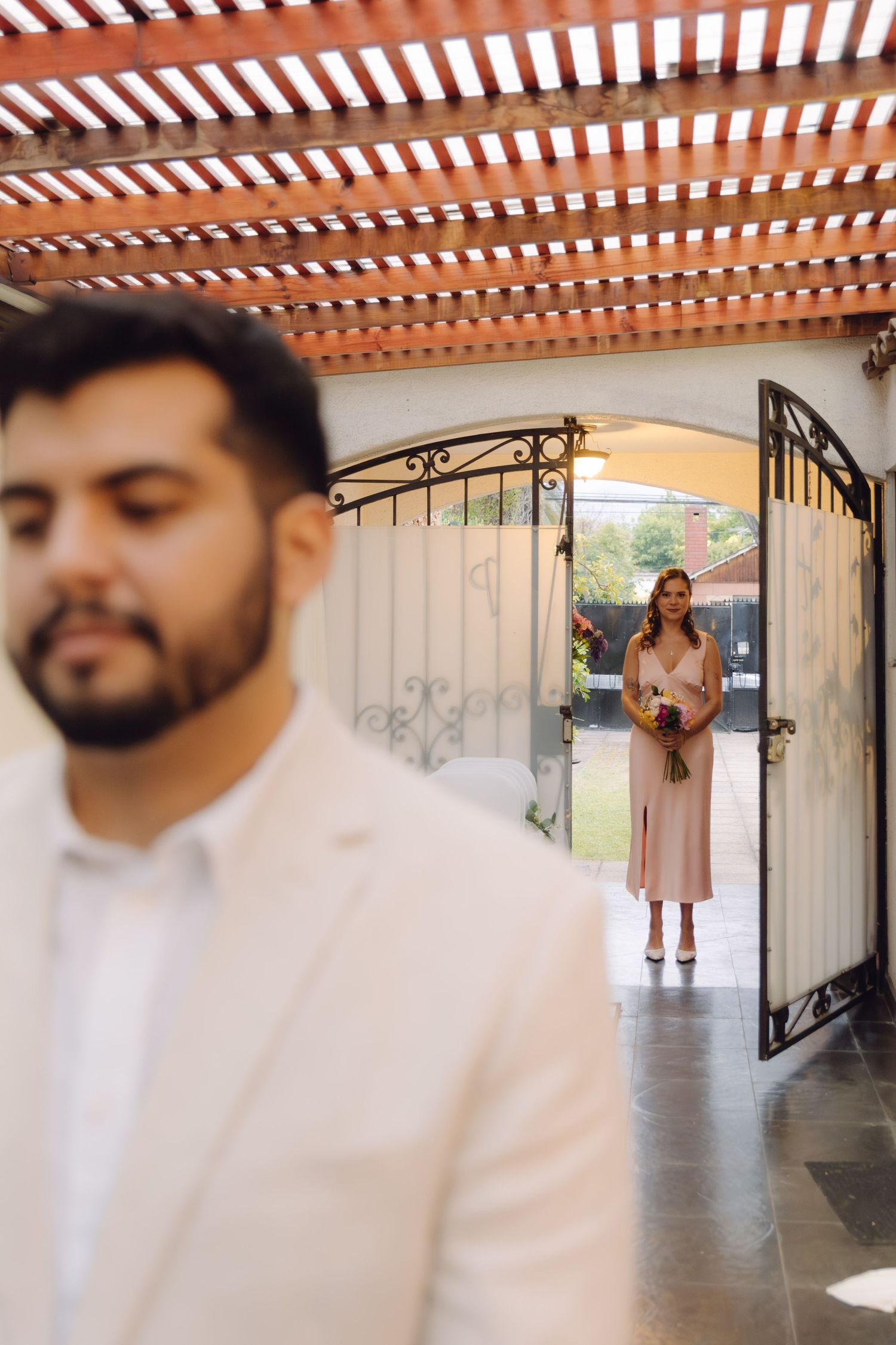 A groom in a white suit stands in the foreground as a bride approaches through garden gates.