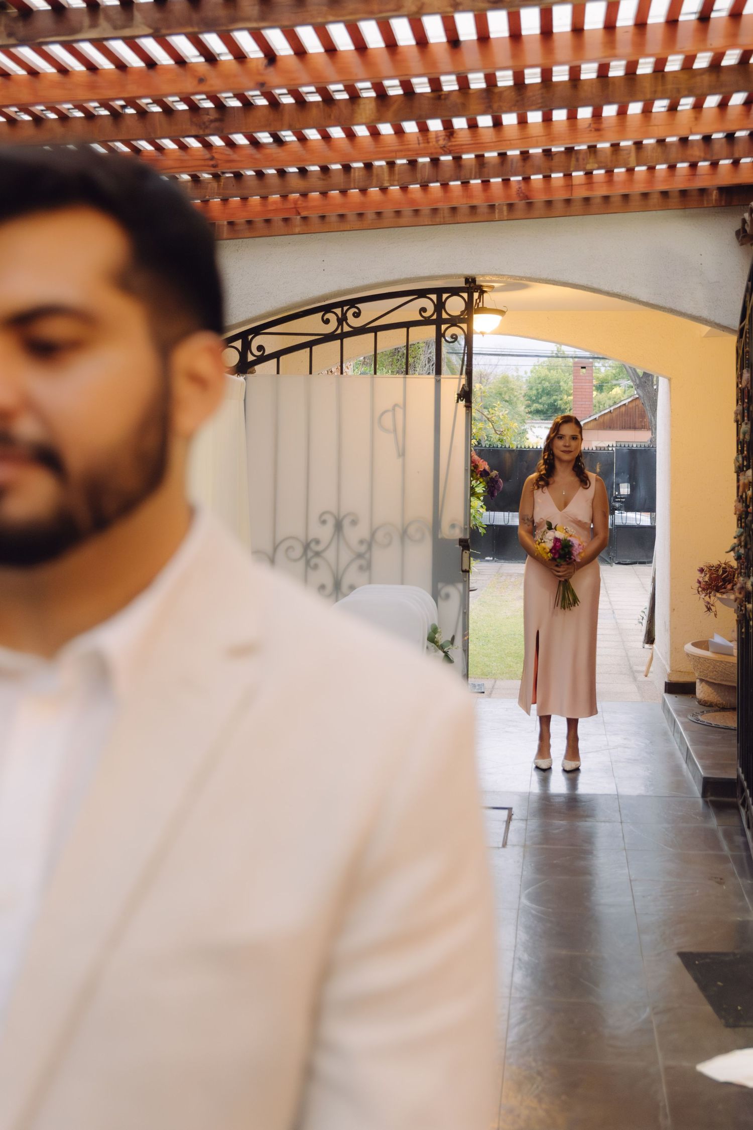 A bride in a pink dress holding a bouquet walks toward a groom under a wooden pergola.