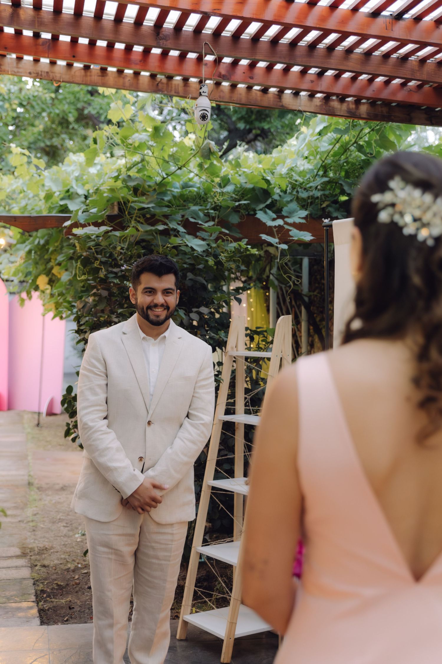 A happy groom in a beige suit smiles at his bride during an outdoor wedding ceremony.