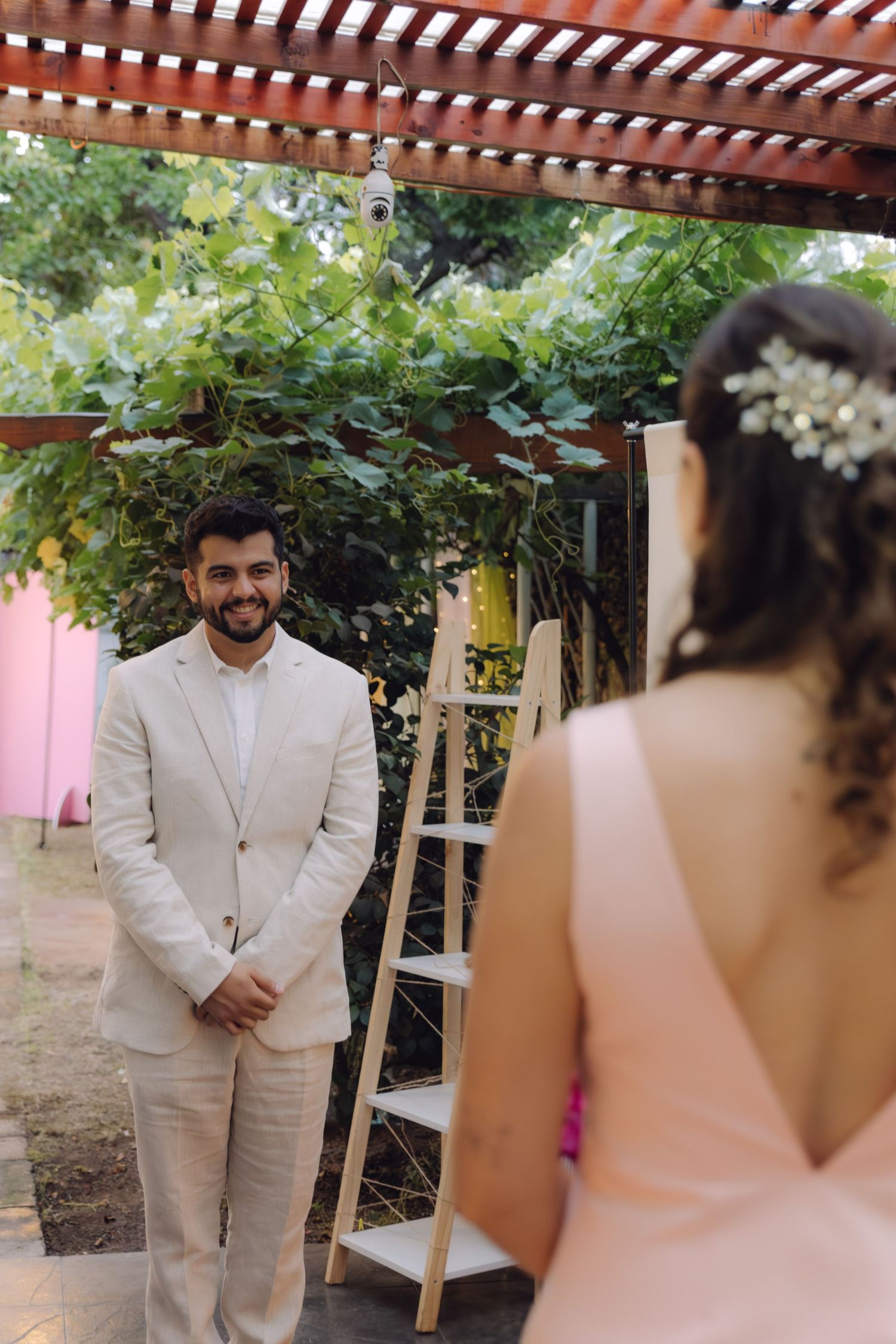 A smiling groom in a beige suit faces his bride outdoors under a lush green pergola.