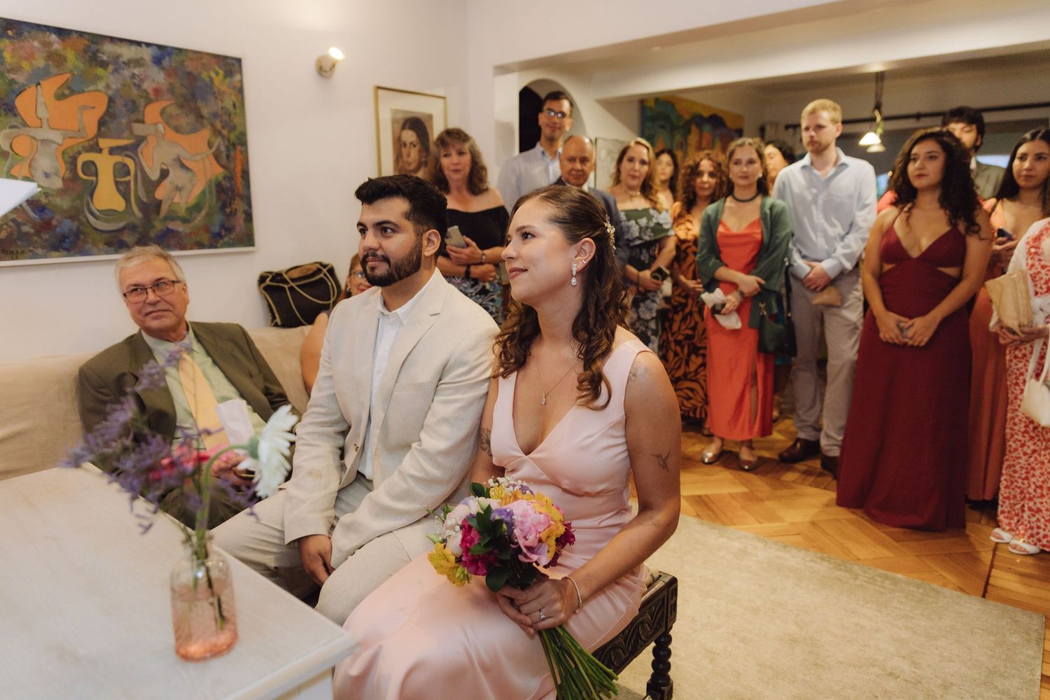 A bride and groom sit together holding a bouquet surrounded by wedding guests indoors.