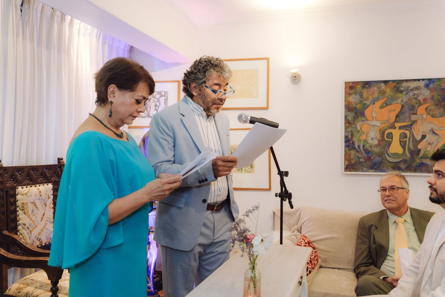 A couple reads vows at a microphone stand during an intimate indoor wedding ceremony while guests watch.