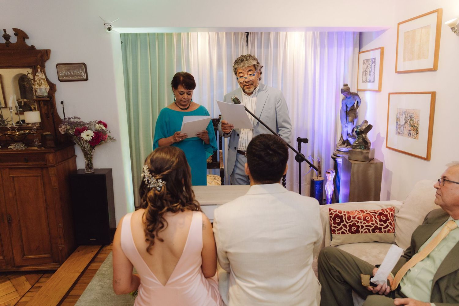 Bride and groom sit before officiants reading vows during a small intimate home wedding ceremony.