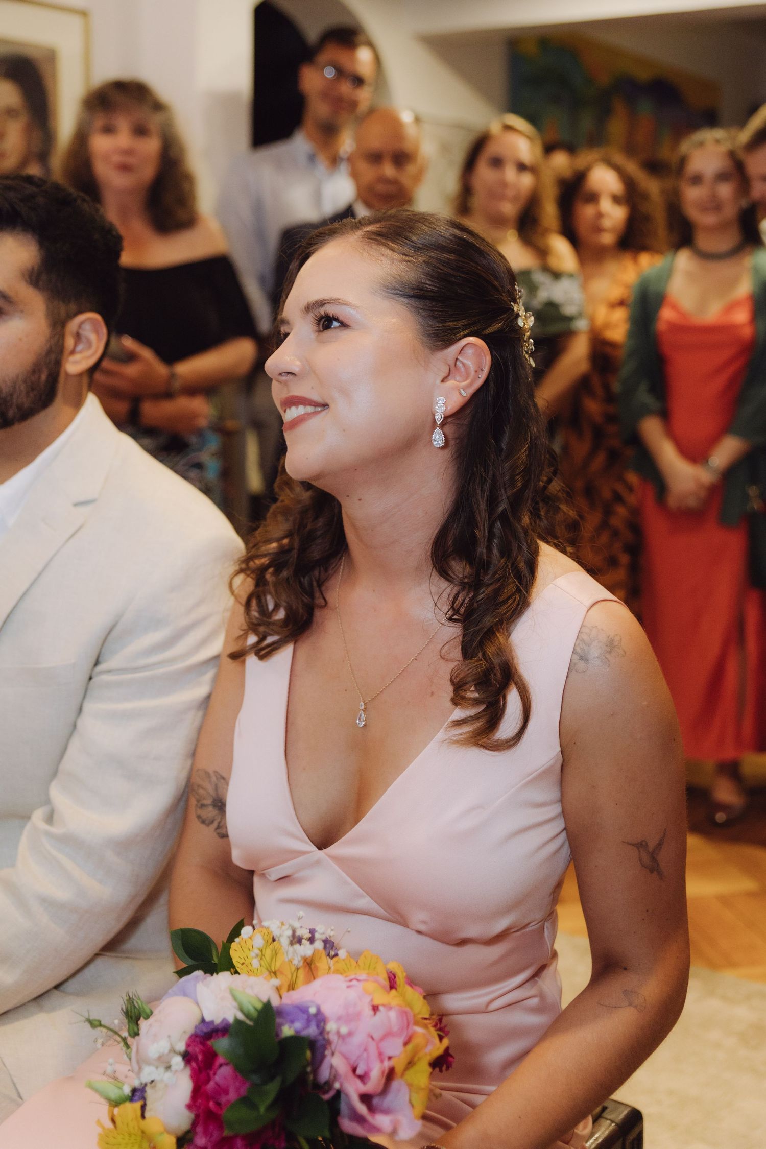Smiling bride in pink dress holds colorful bouquet during intimate indoor wedding ceremony with guests.