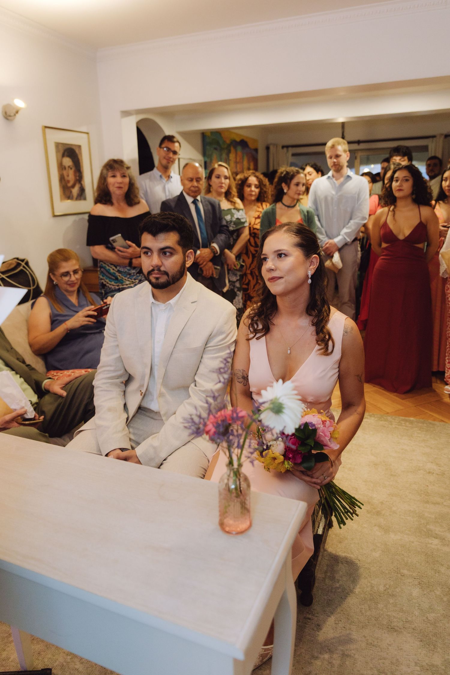 Bride holding colorful bouquet sits beside groom in white suit during intimate indoor wedding ceremony.