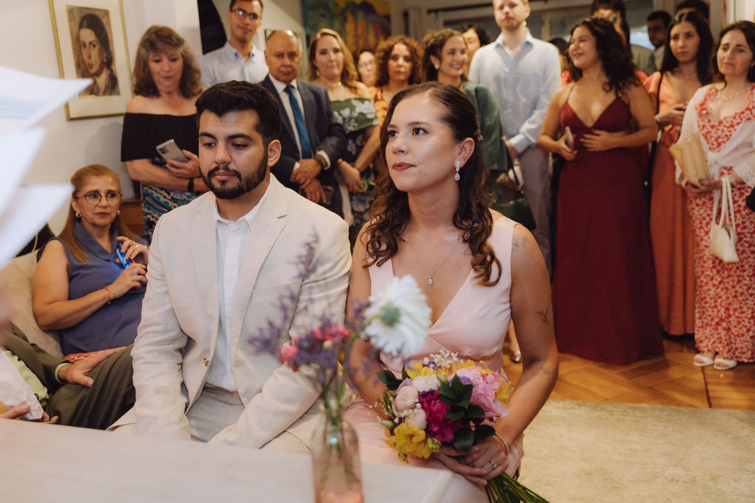Bride and groom sit together at table during intimate indoor wedding ceremony surrounded by guests.