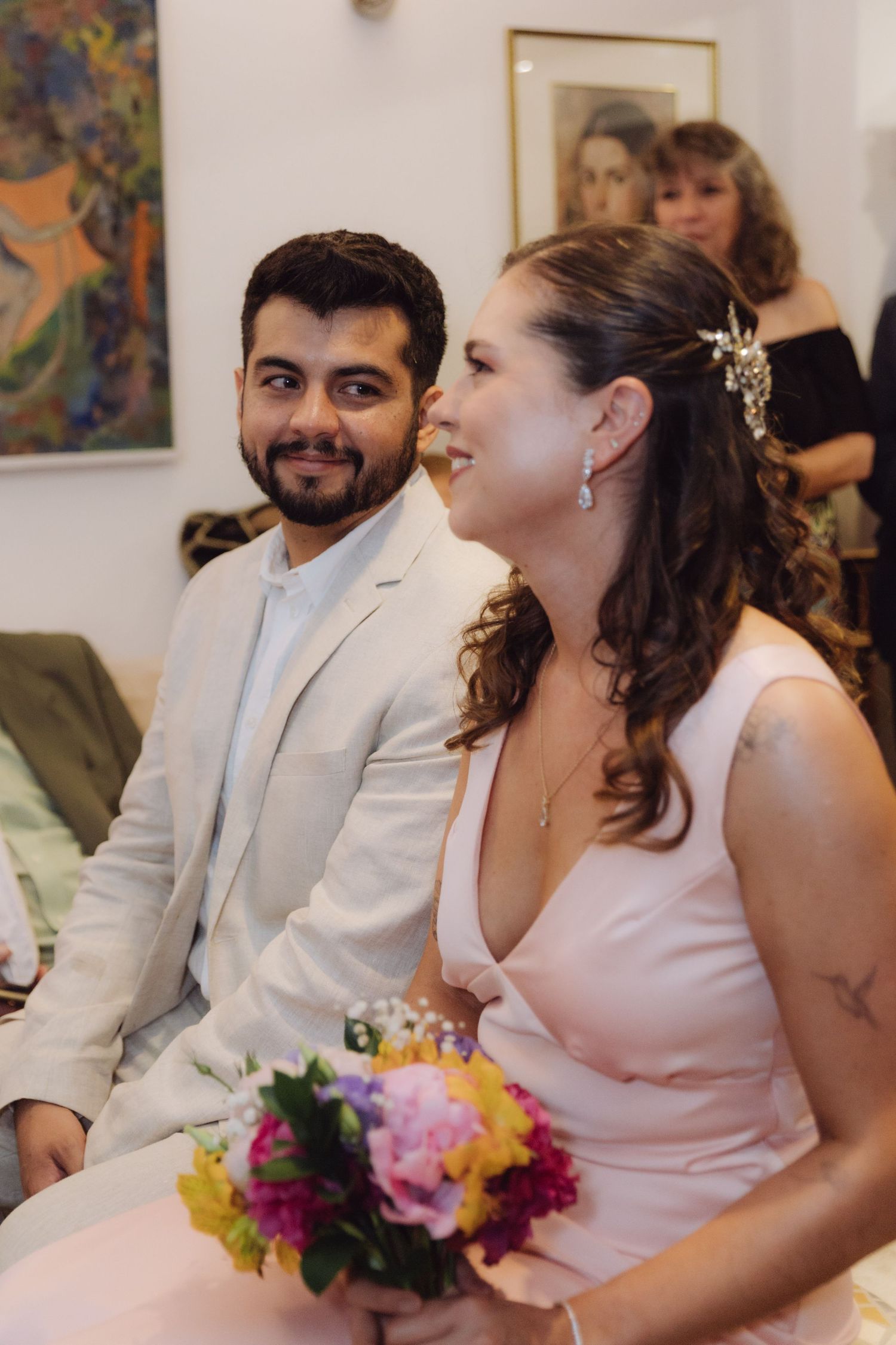 Groom in white suit and bride holding colorful bouquet seated together during wedding ceremony.