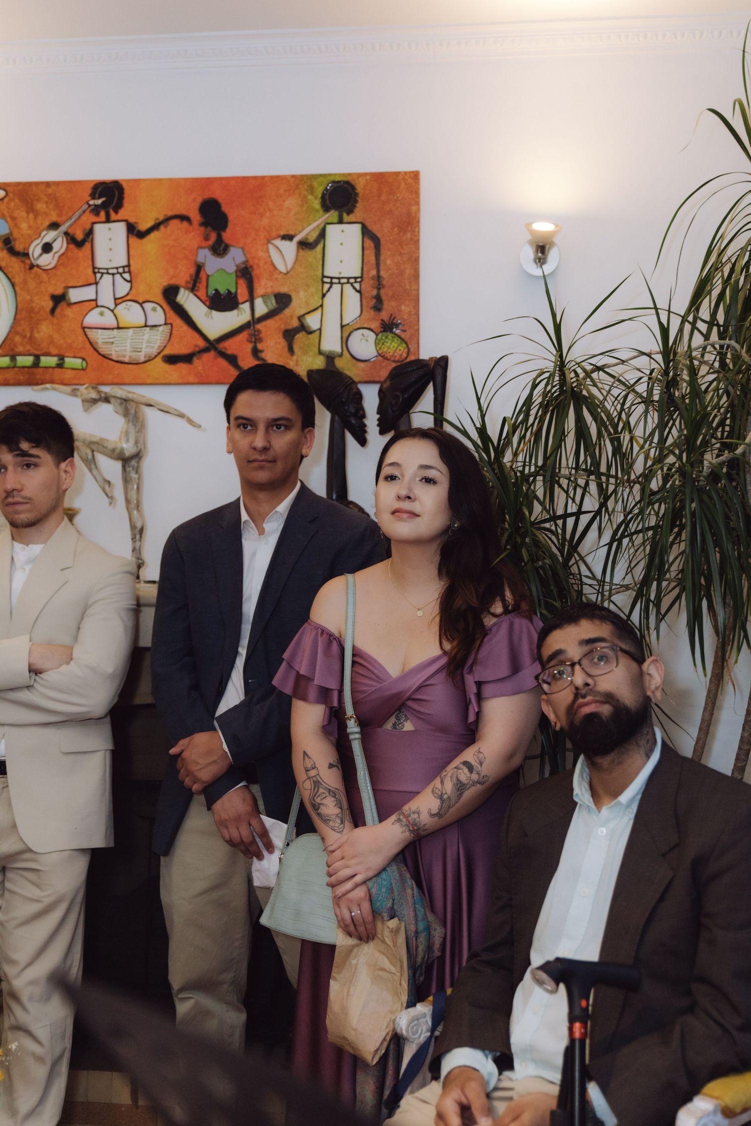 Wedding guests in formal attire gathered indoors near colorful artwork and tropical plant decor.