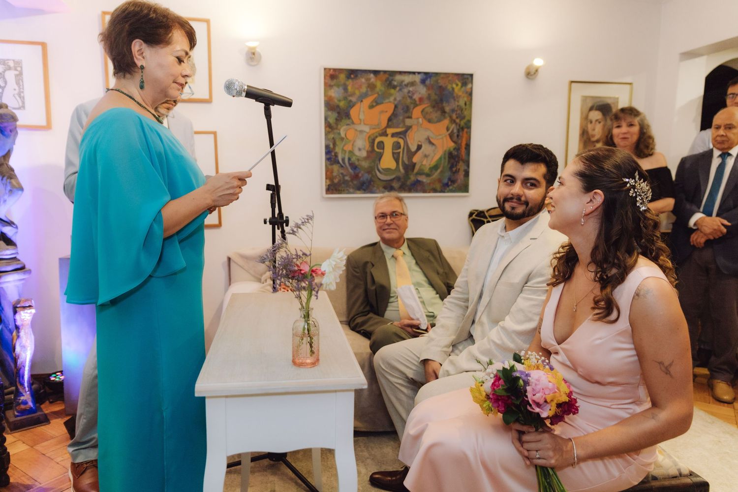 Woman in teal dress speaks at microphone during intimate indoor wedding ceremony with smiling couple seated nearby.