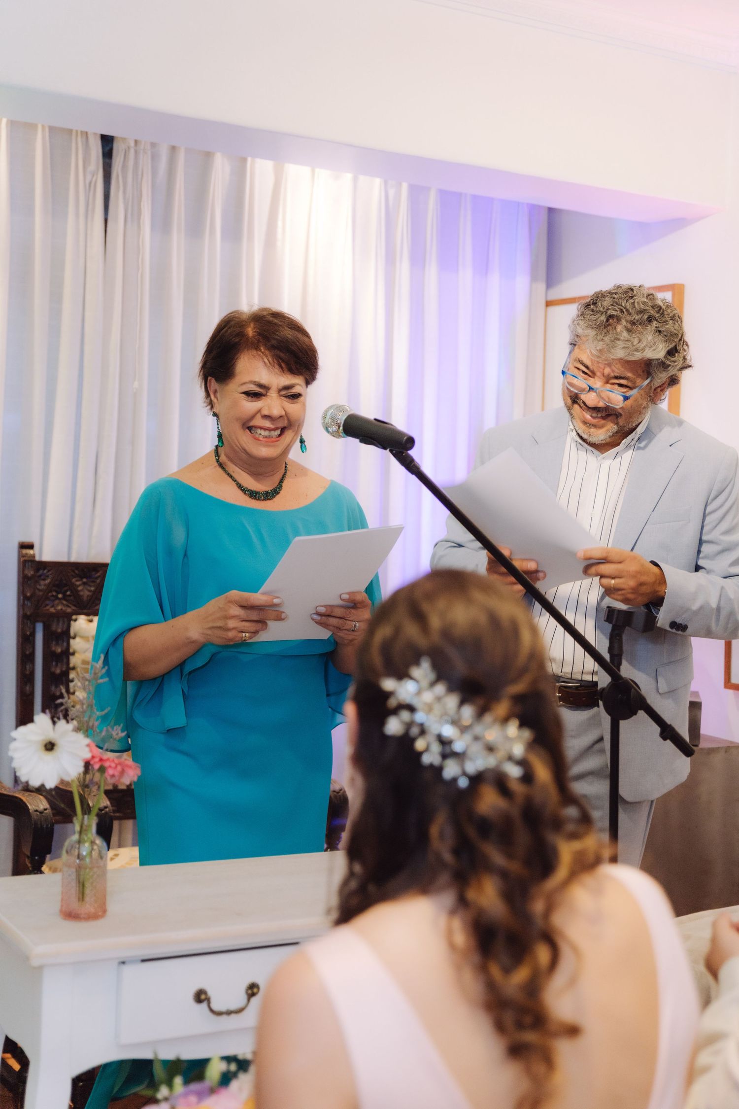 Smiling woman in teal dress reads from paper at microphone during wedding ceremony beside man in gray suit.
