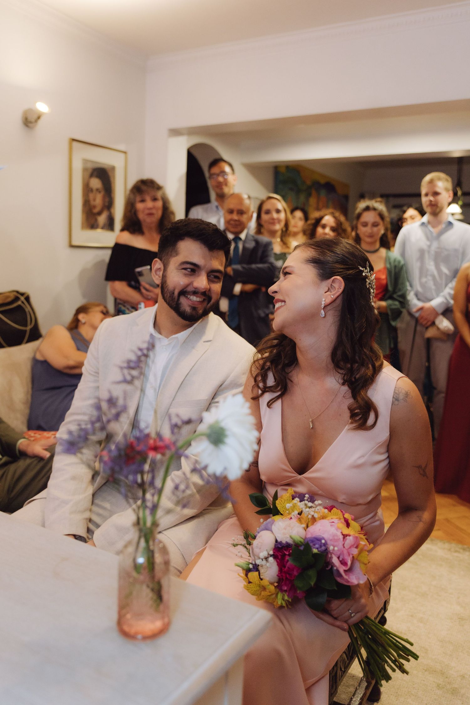 Happy bride holding colorful bouquet and groom in white suit smile during indoor wedding ceremony with guests.