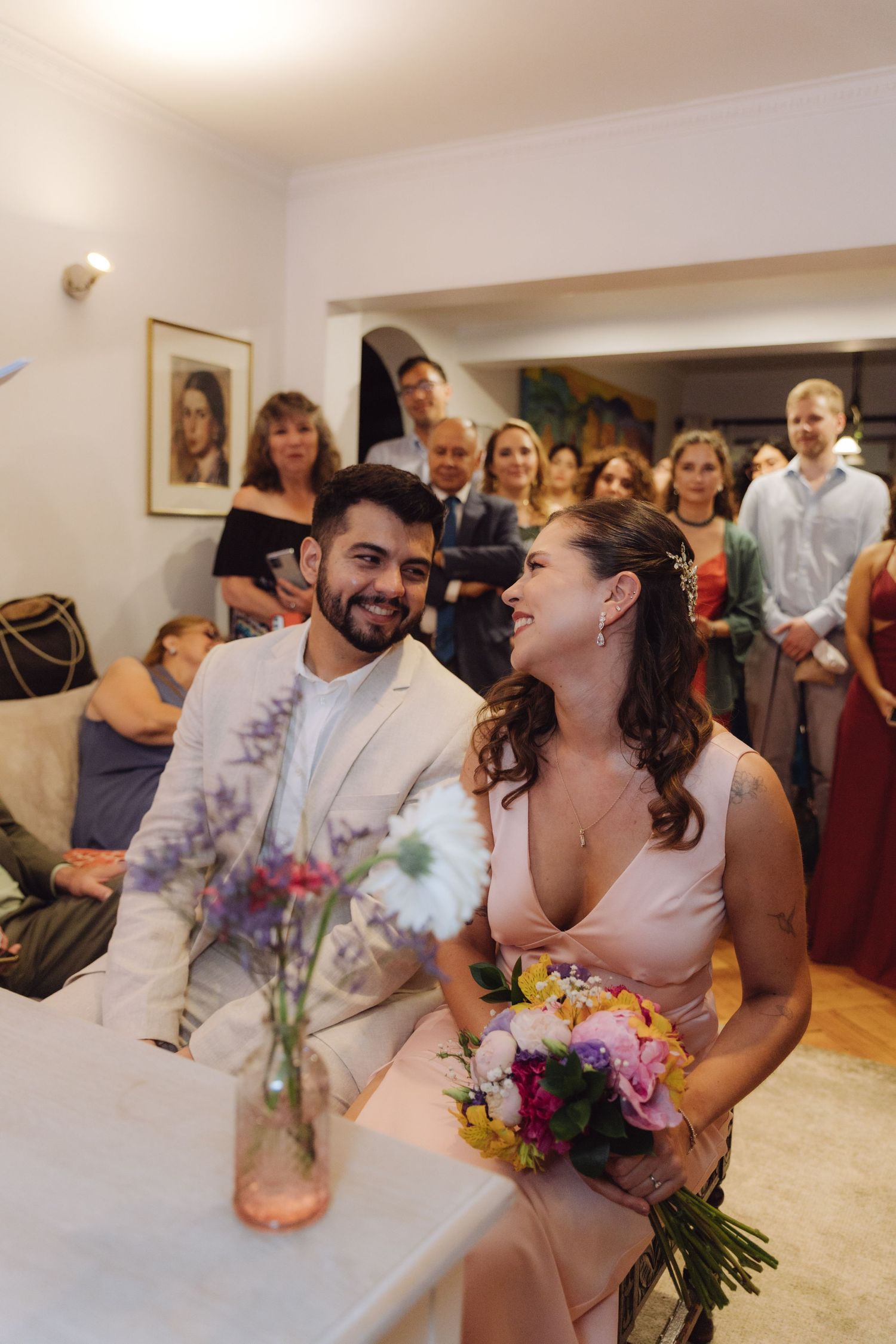Bride with floral bouquet and groom in white suit share a joyful moment during intimate home wedding ceremony.