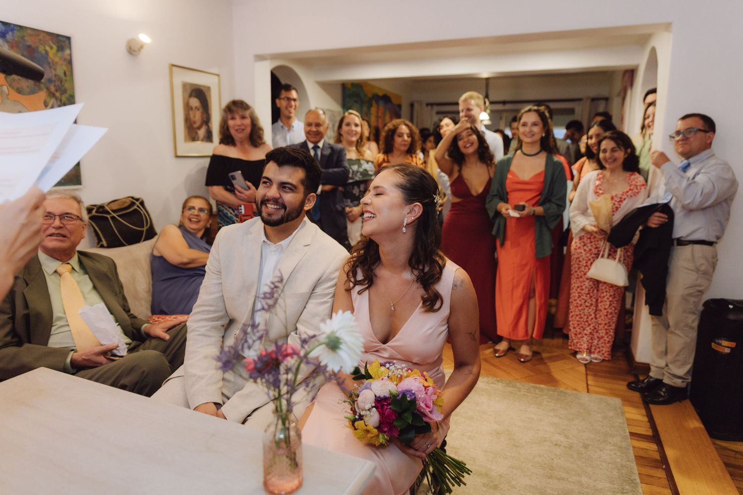 Smiling bride holding colorful bouquet sits with groom during indoor wedding ceremony surrounded by standing guests.