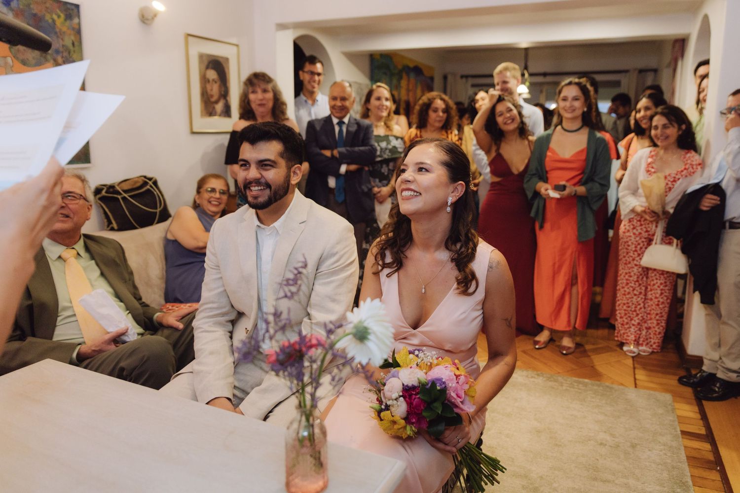 Bride and groom smile joyfully during intimate indoor wedding ceremony with colorful floral arrangements and guests behind.