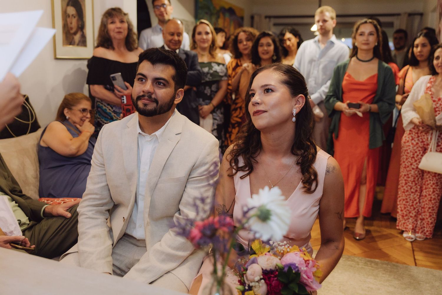 Bride in pink dress and groom in beige suit sit attentively during cozy indoor wedding ceremony with guests.