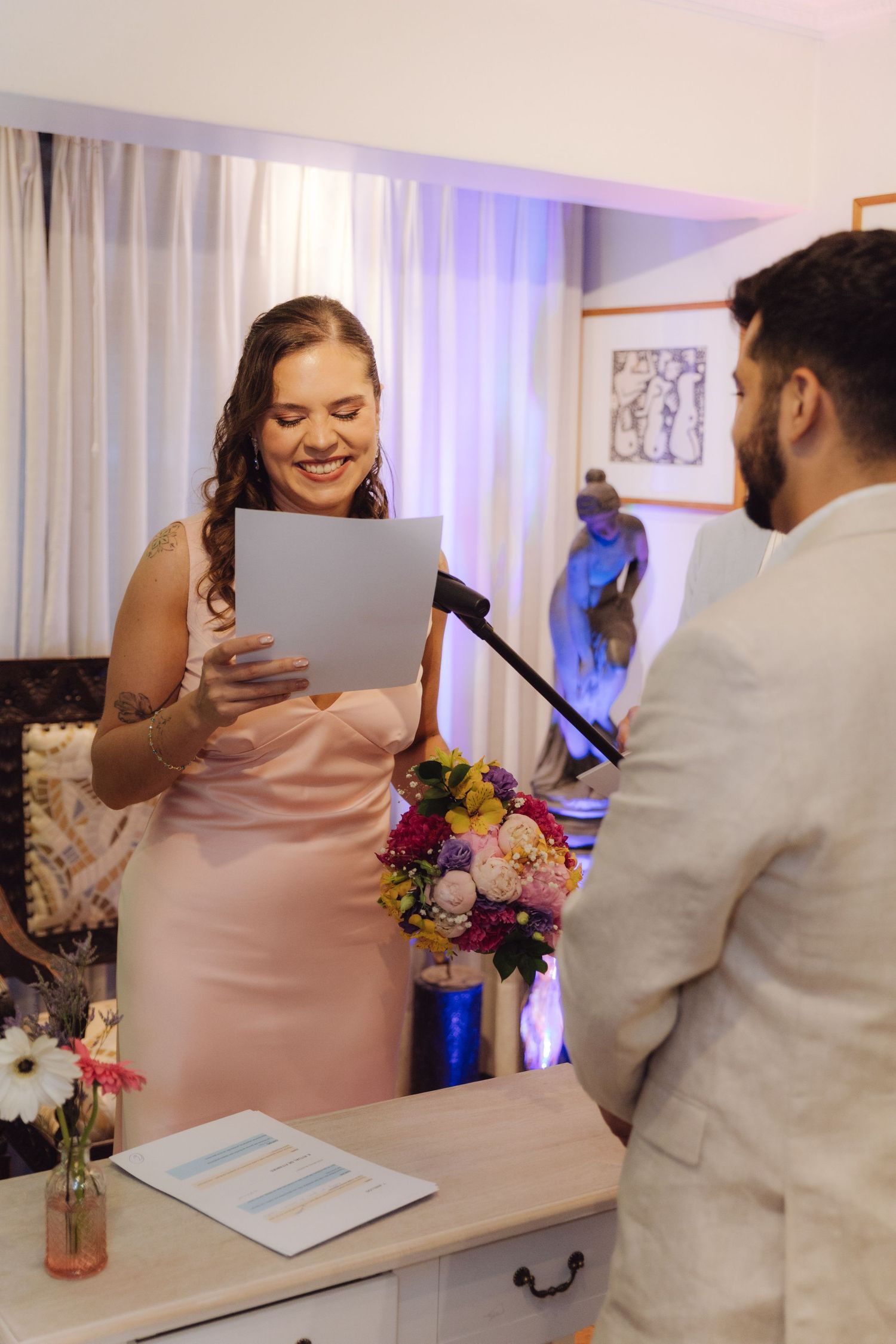 Radiant bride in pink dress reads wedding vows from paper at microphone facing groom in white suit.