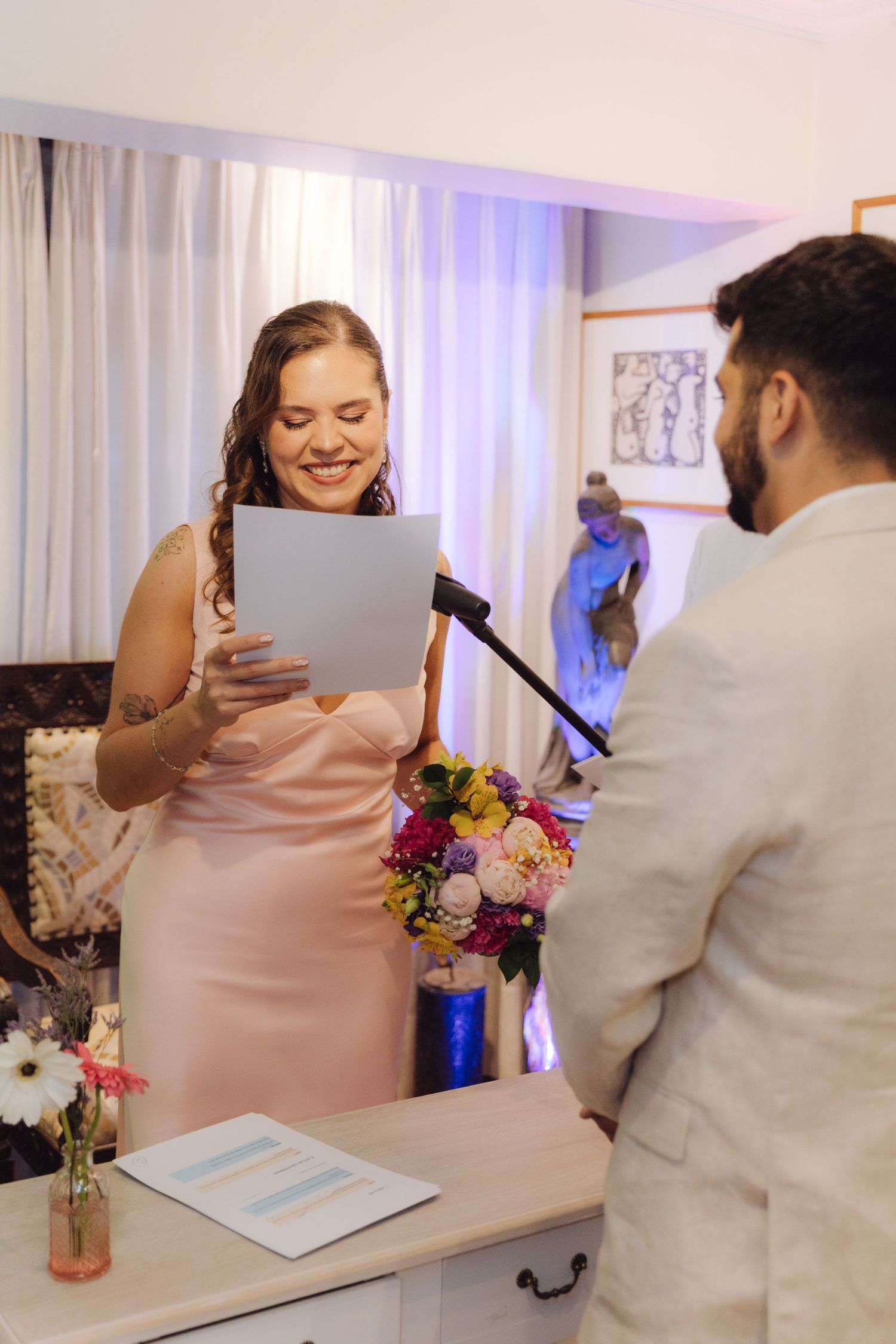Smiling bride in pink gown reads heartfelt vows from paper to groom during intimate indoor wedding ceremony.
