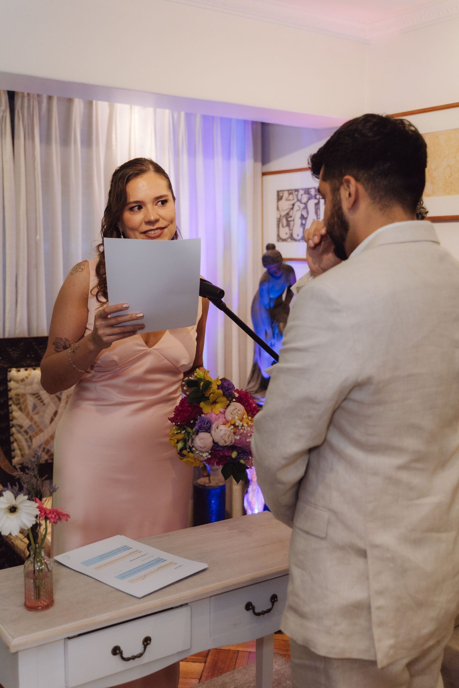 Bride in blush pink dress reads wedding vows to groom at indoor ceremony with floral decorations nearby.