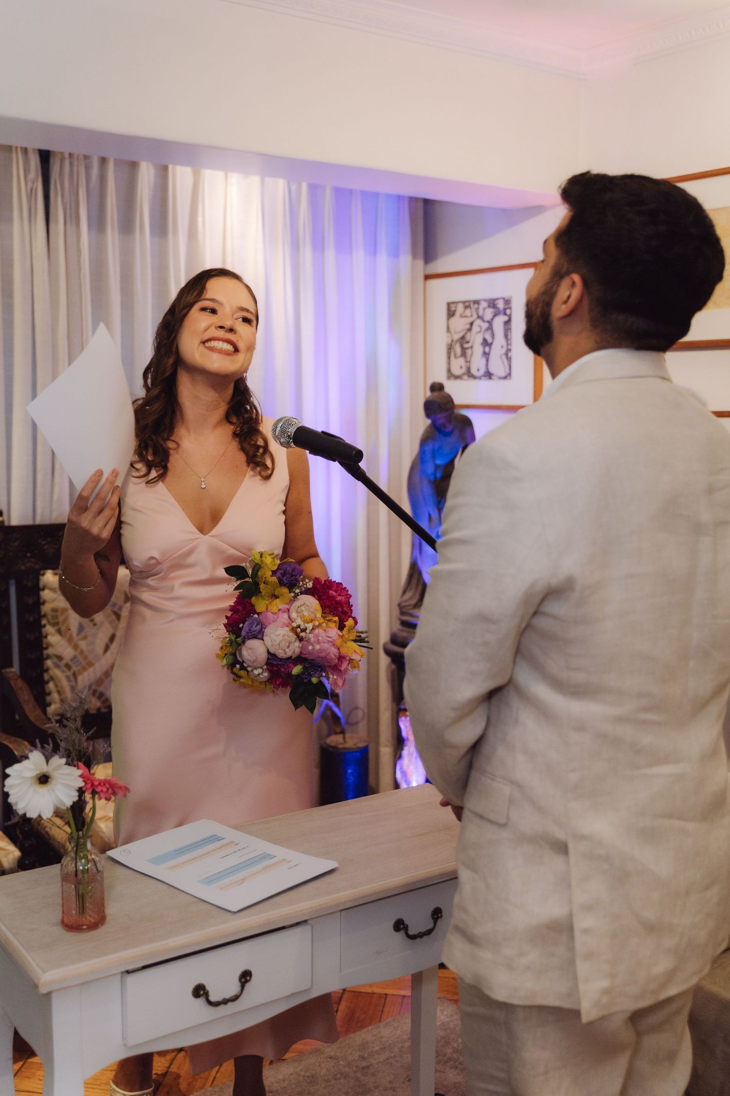 Bride in pink dress reads vows at microphone stand during intimate indoor wedding ceremony.