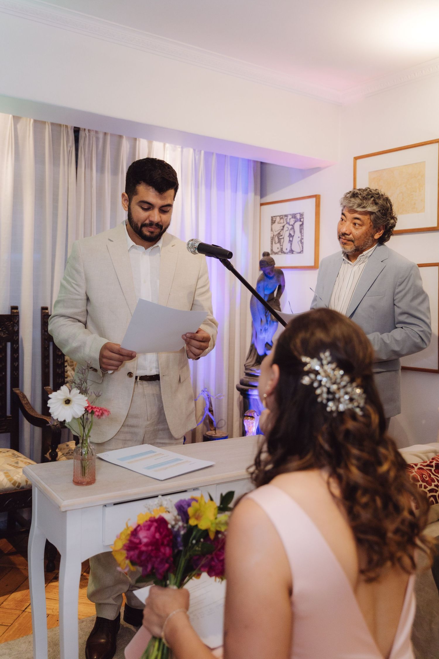 Groom in beige suit reads wedding vows while officiant stands behind him at indoor ceremony.