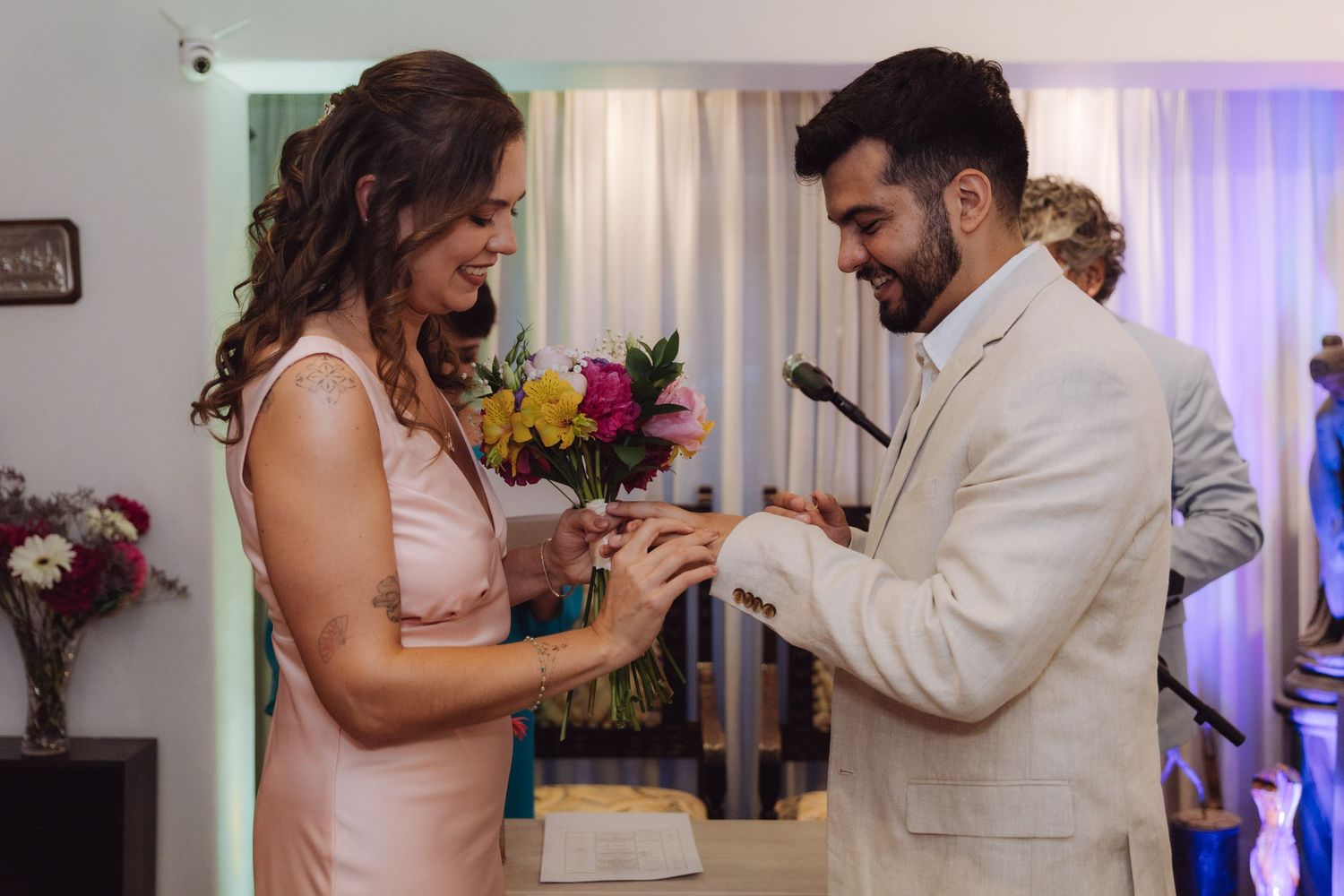 Couple exchanges rings during intimate indoor wedding ceremony, both smiling happily.