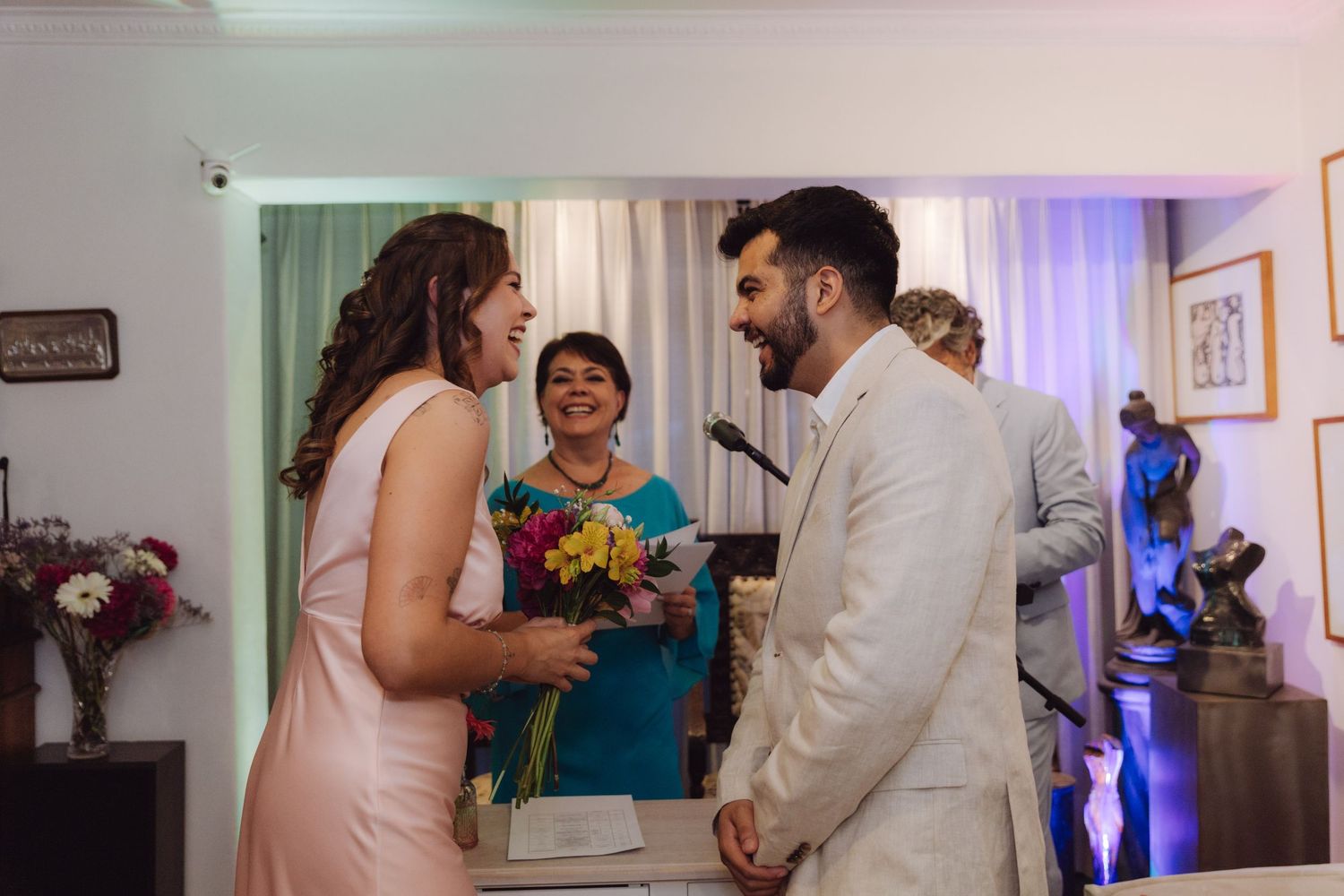 Bride and groom share a joyful moment with officiant during small indoor wedding ceremony.