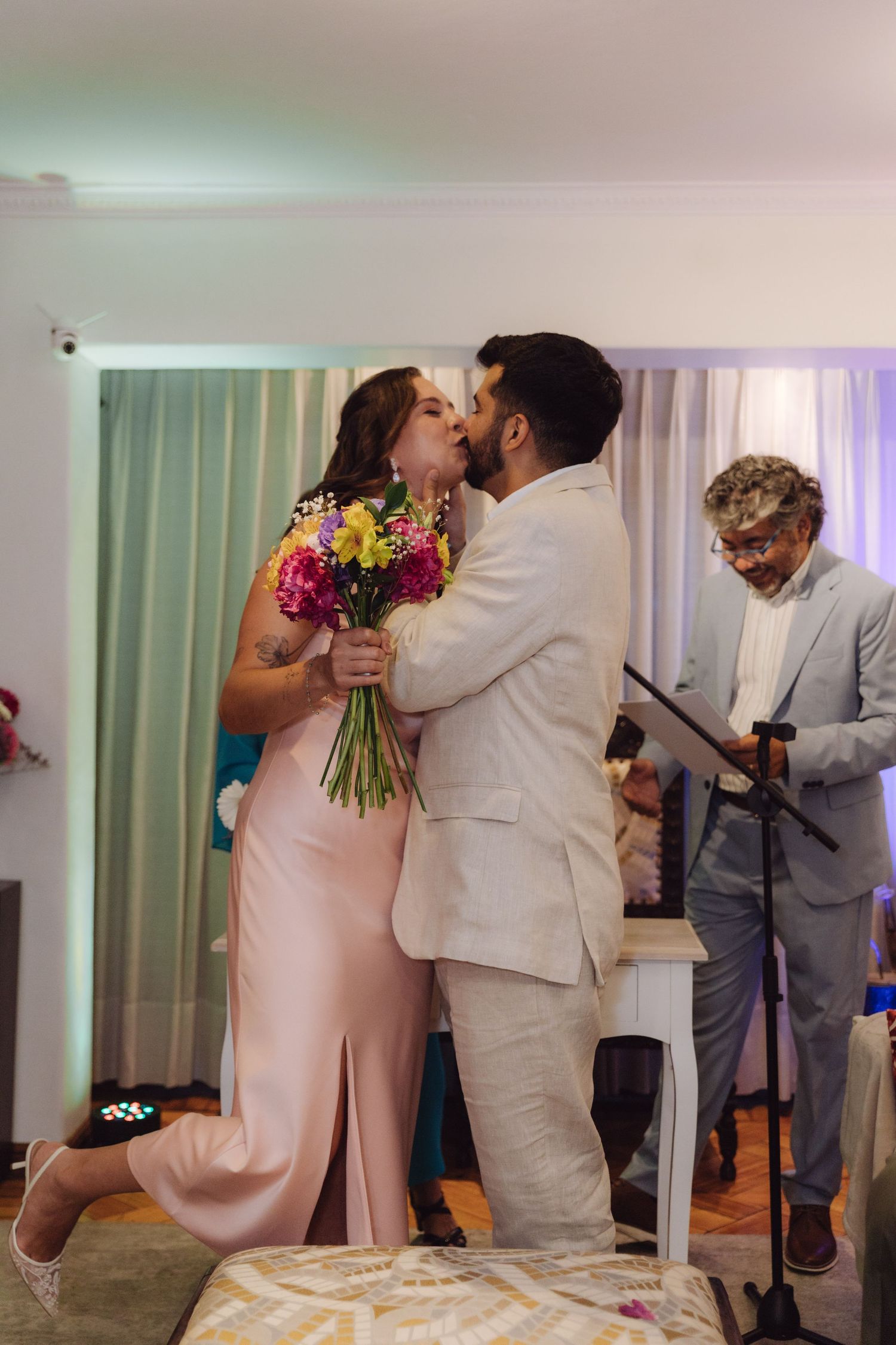 Bride and groom share first kiss holding colorful bouquet during indoor wedding ceremony.