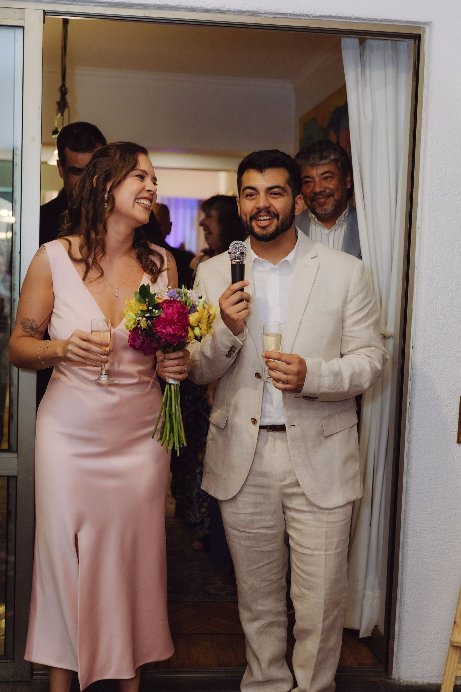 Newlyweds smile holding bouquet and champagne glasses while groom speaks into microphone.